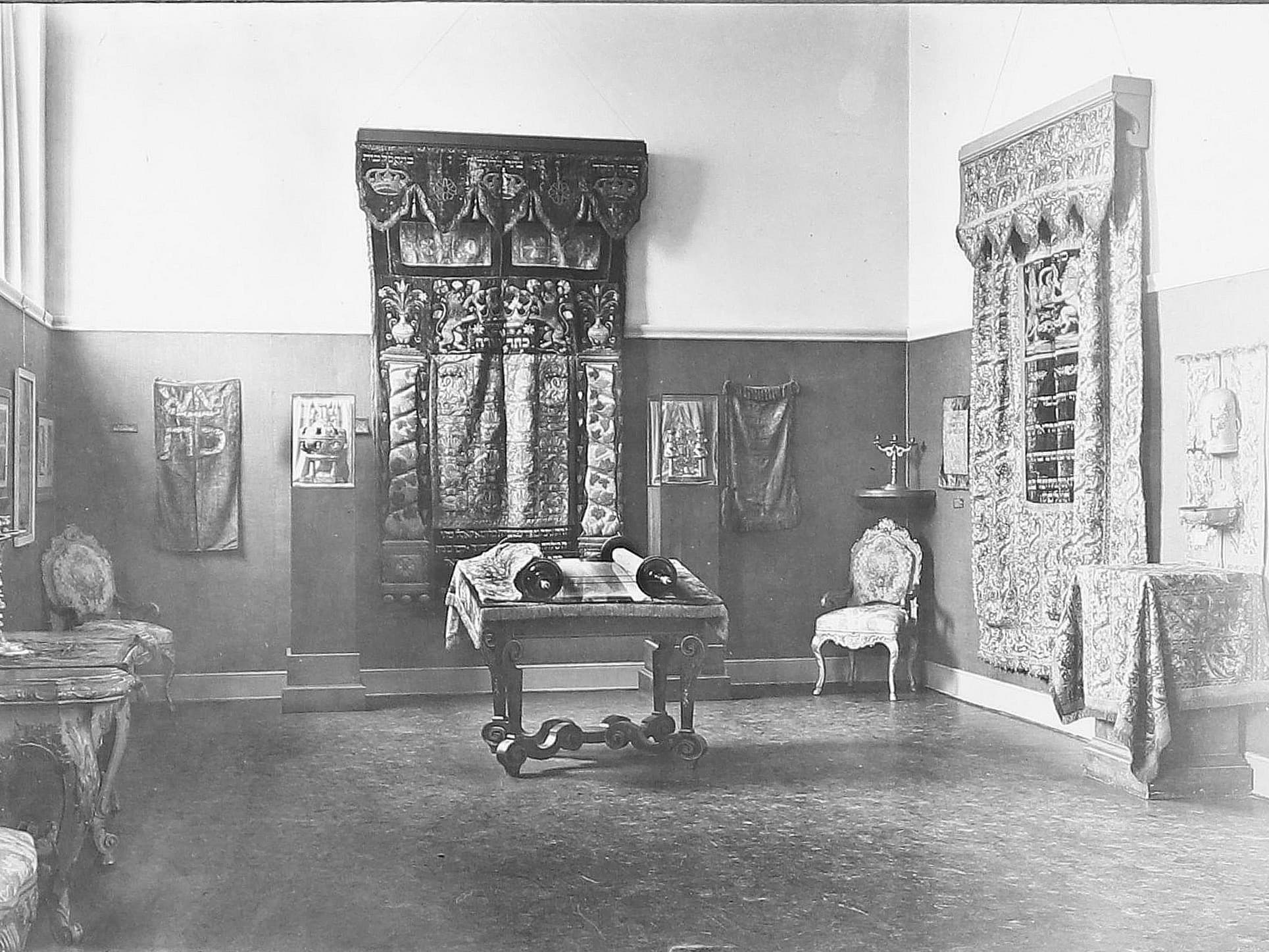 Ein historischer Raum mit kunstvollen Wandteppichen, einem Holztisch mit Schriftrollen und Vintage-Stühlen. An den Wänden ist kunstvolle Textilkunst zu sehen.