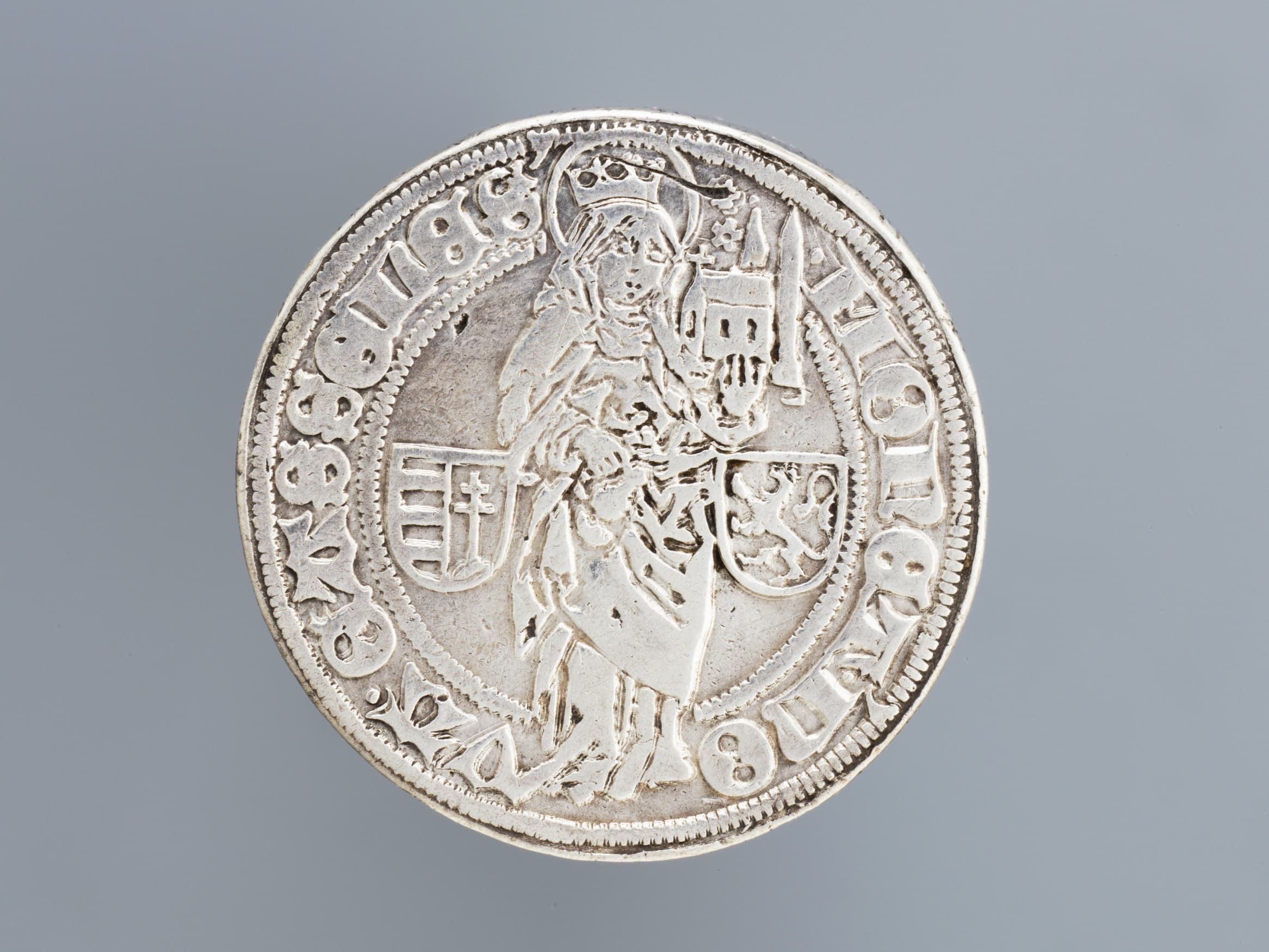 Silbermünze mit einer gekrönten Figur, die ein Gebäude hält, umgeben von dekorativem Text und zwei Schildern.