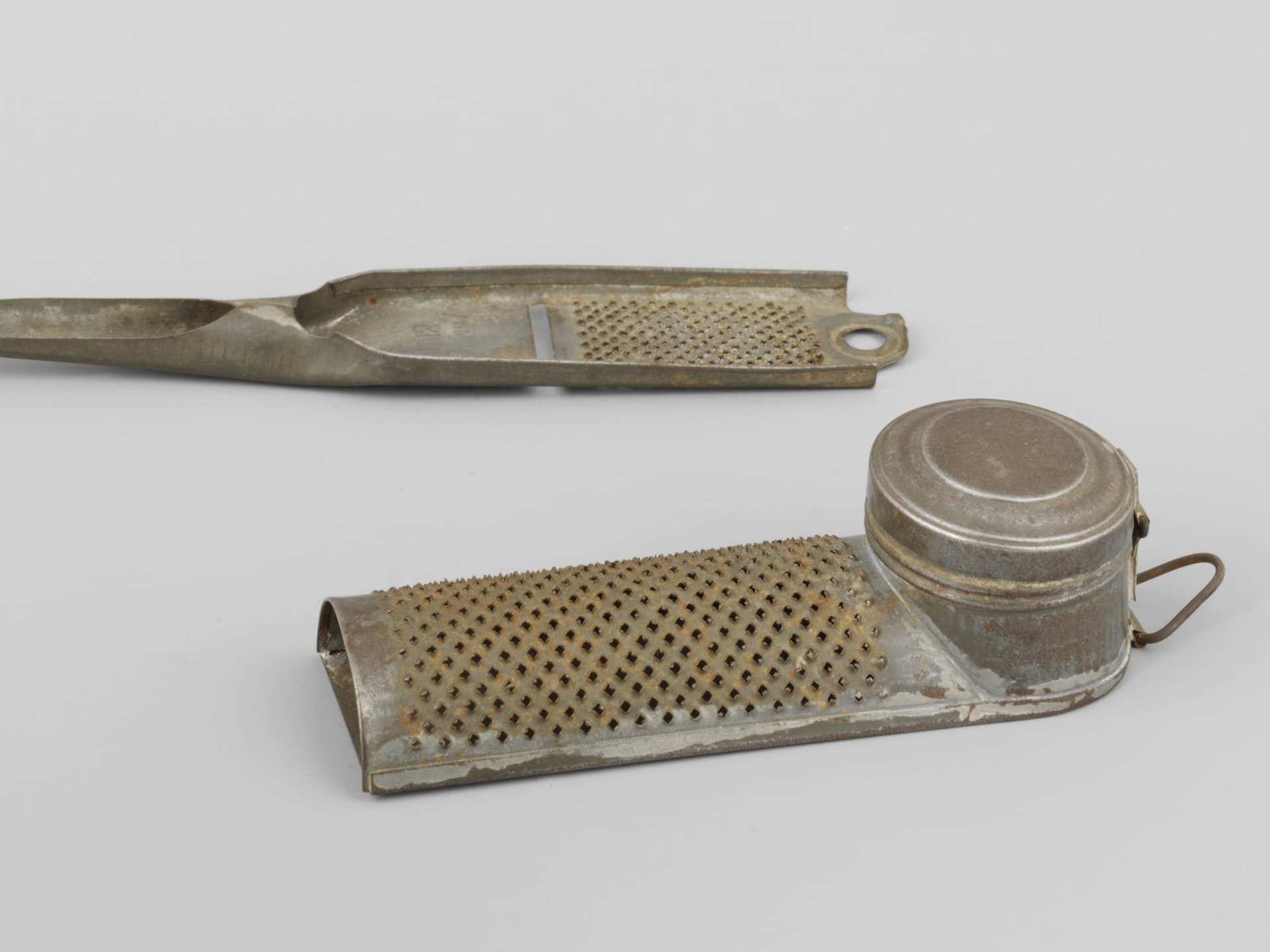 Two vintage metal graters on a plain background; one is flat with a handle, the other is cylindrical with a lid.