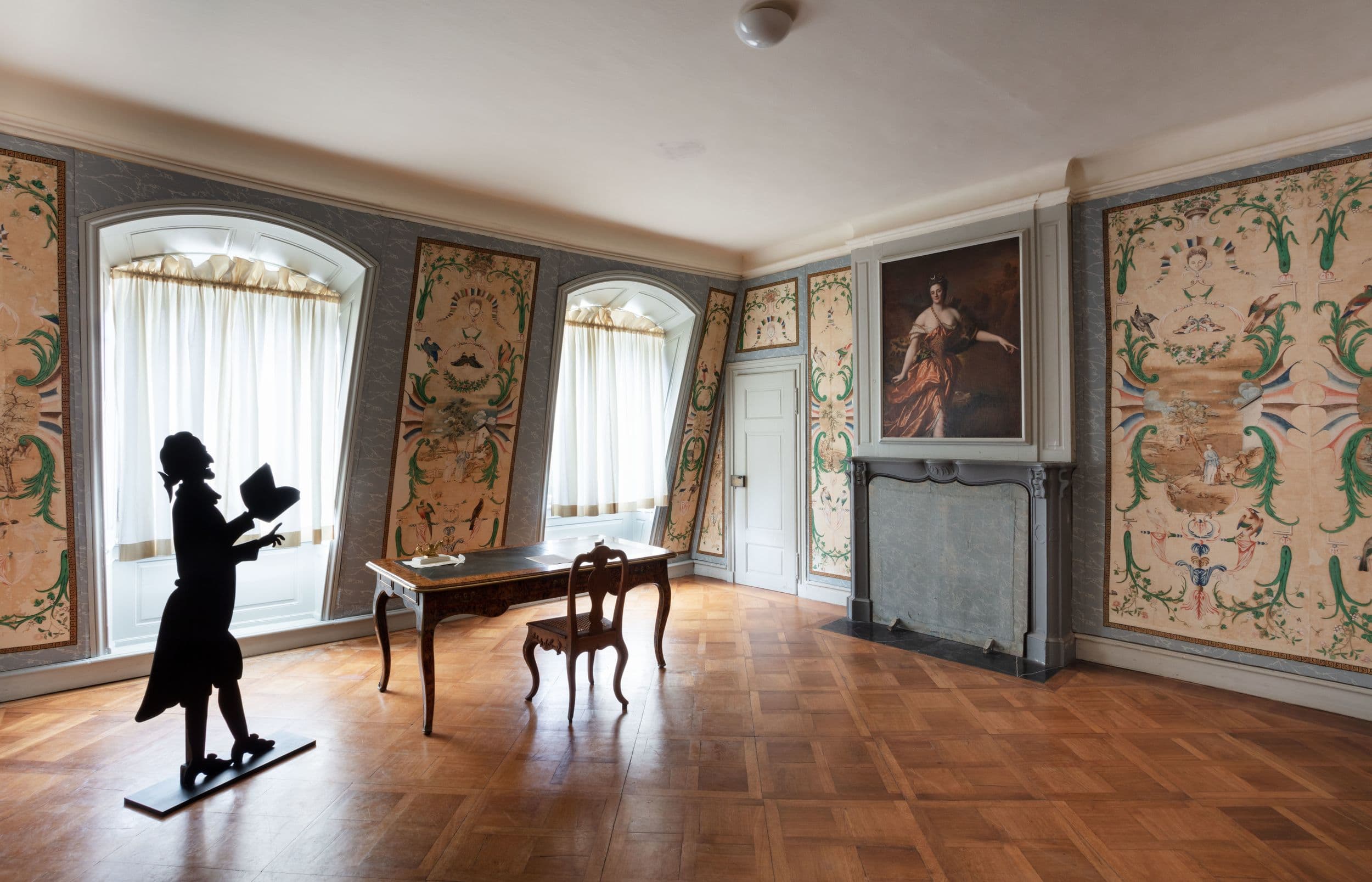 Eleganter Raum mit kunstvoller Tapete, einem Holztisch, Stühlen, einer Silhouettenskulptur und einem großen Porträt über einem Kamin.