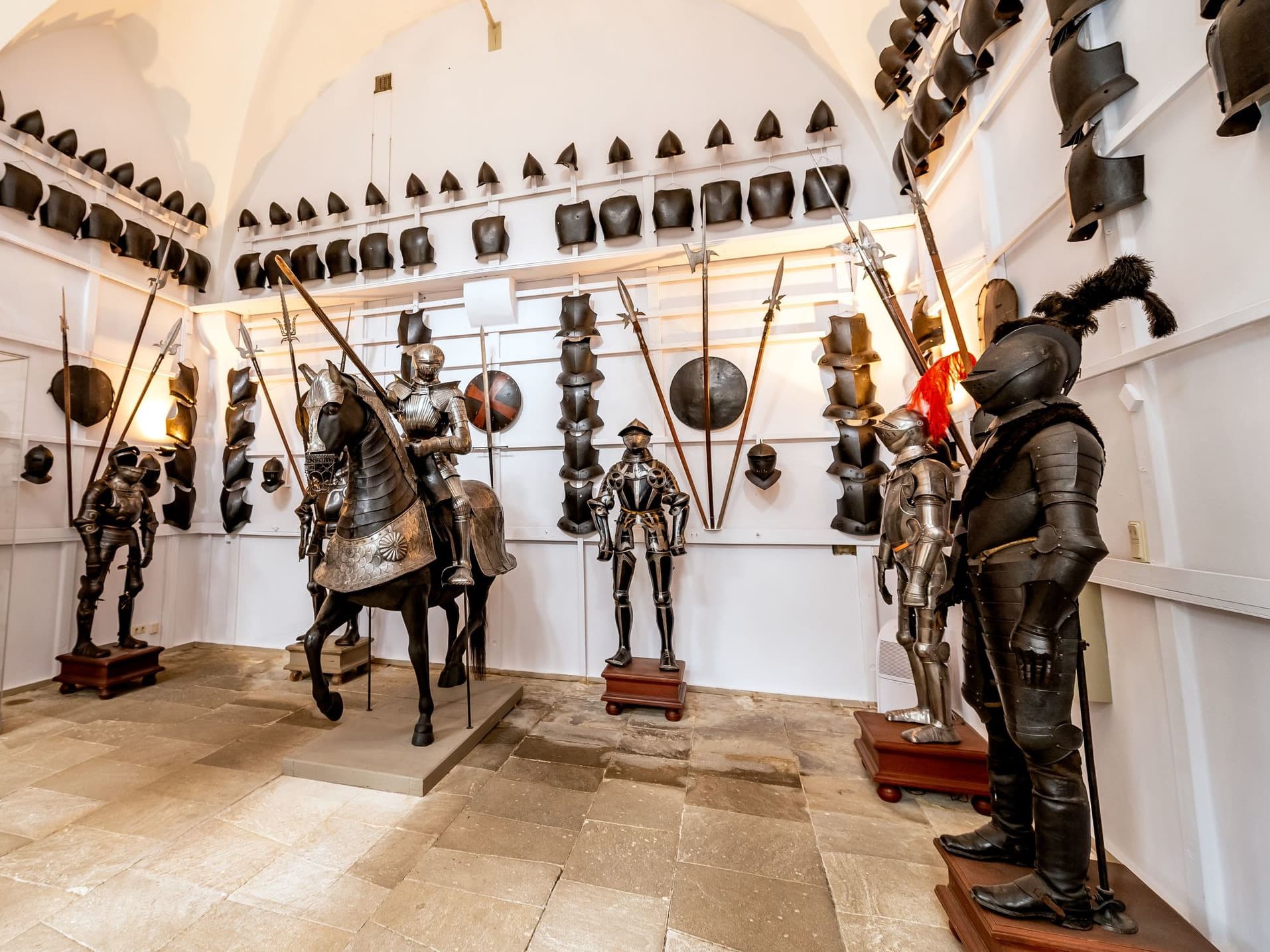 Ein mittelalterlicher Waffenraum mit Rüstungen, darunter ein Ritter auf einem Pferd, umgeben von Helmen und Schilden an weißen Wänden.