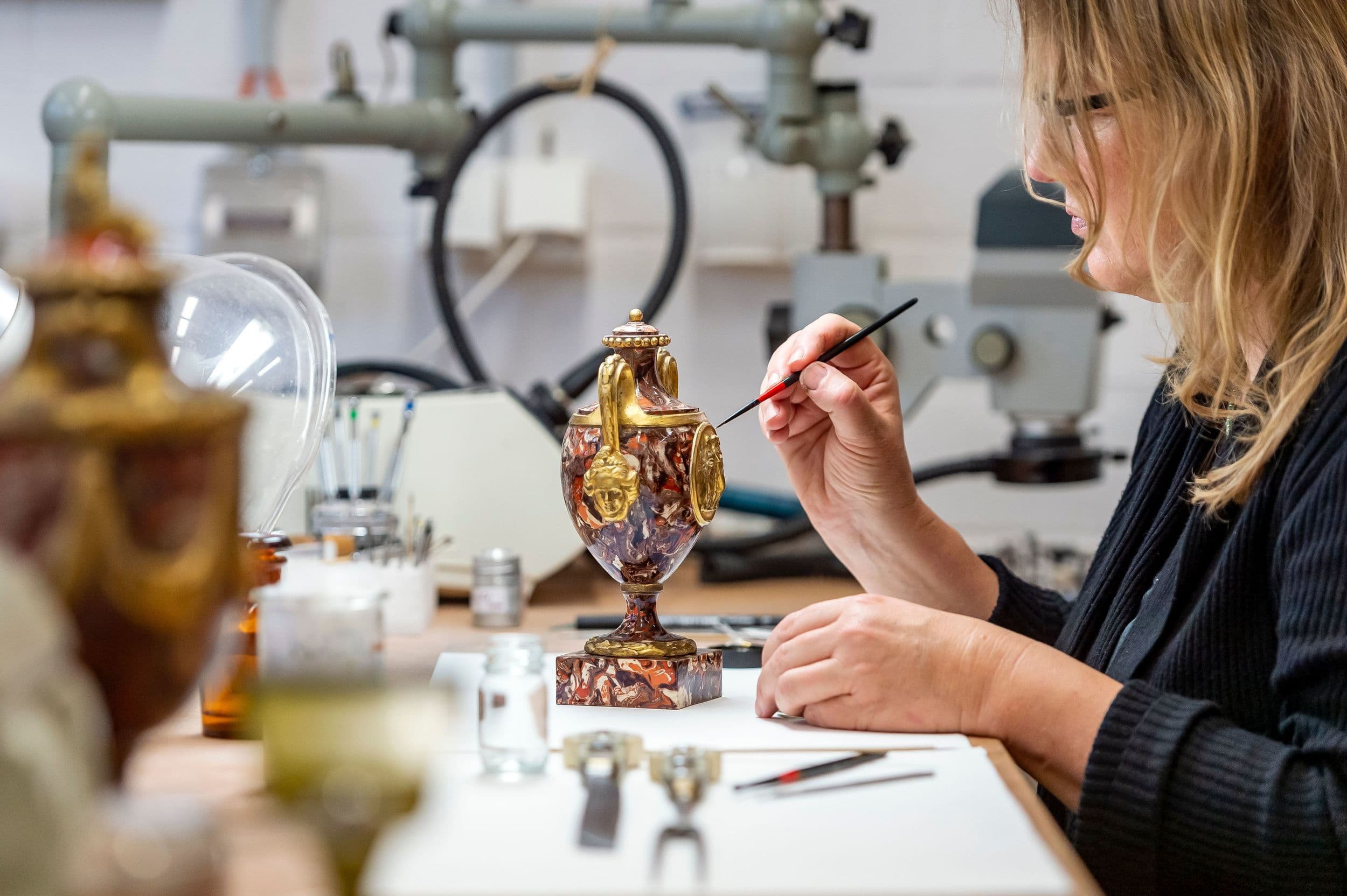 Eine Frau restauriert in einer Werkstatt, umgeben von Werkzeugen und Geräten, sorgfältig eine kunstvolle Vase.