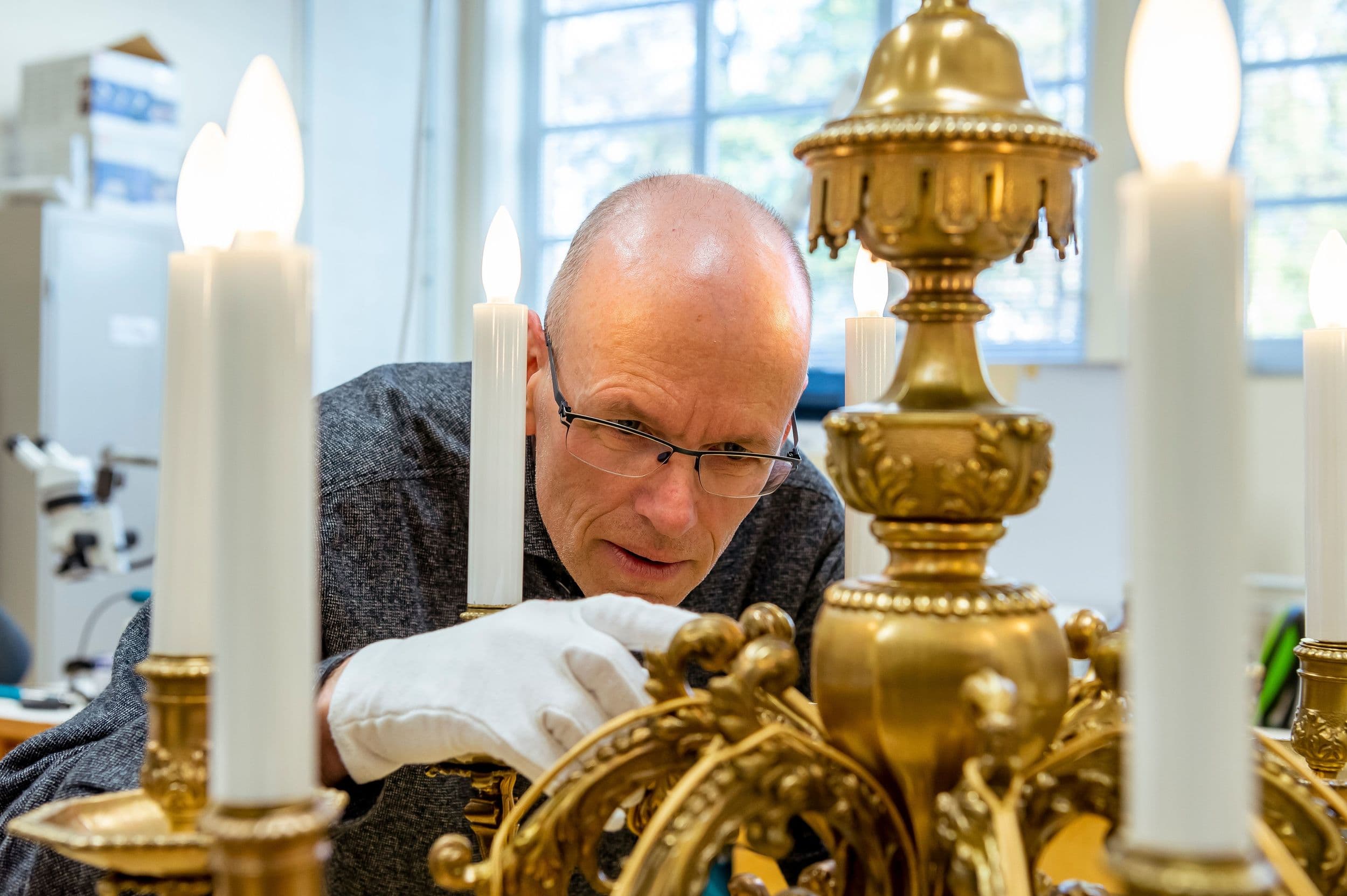 Eine Person mit Handschuhen untersucht sorgfältig einen kunstvollen, goldfarbenen Kronleuchter mit brennenden kerzenförmigen Glühbirnen in einem gut beleuchteten Raum.