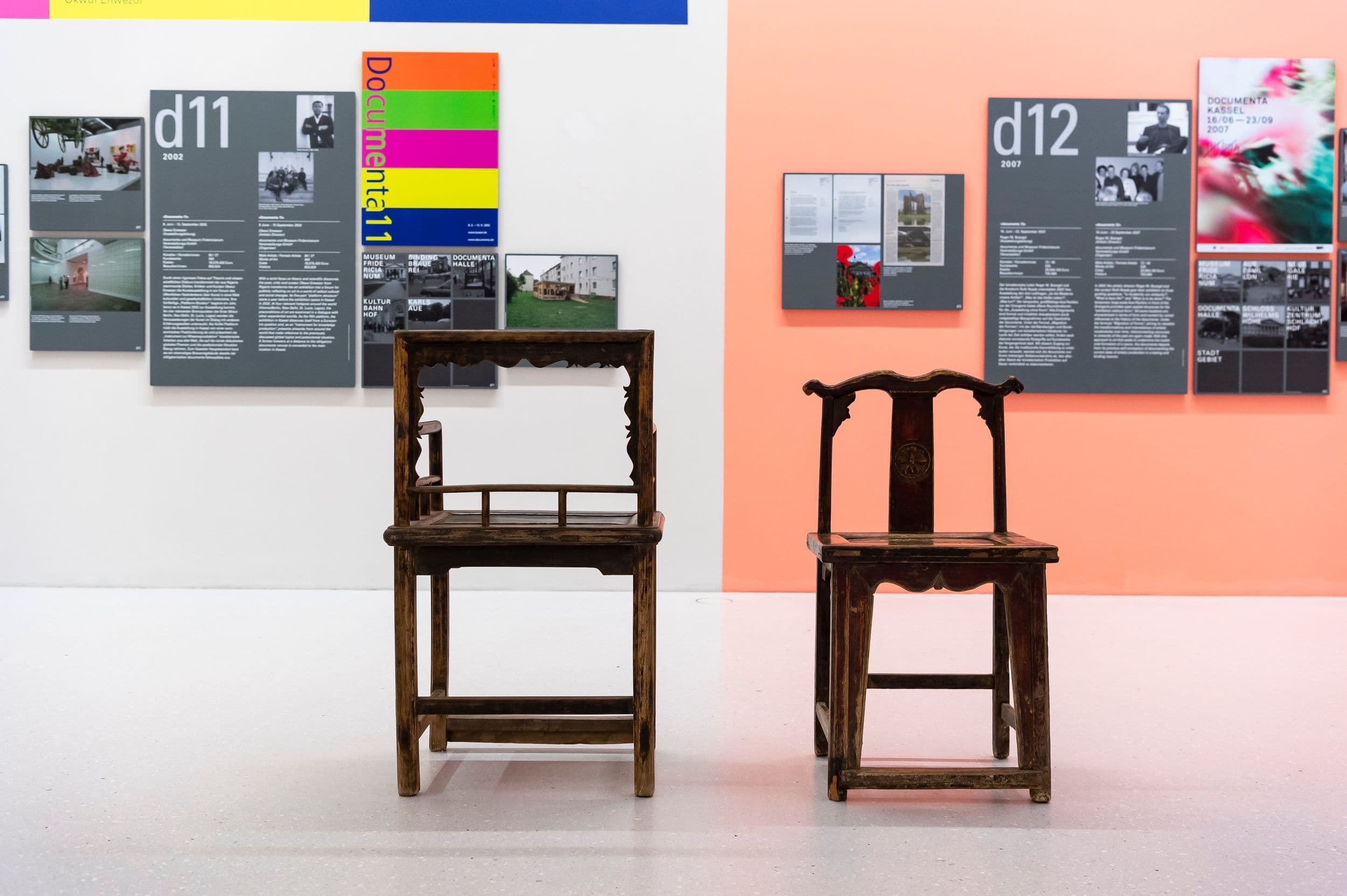 Zwei Holzstühle in einer Galerie mit farbenfrohen Wanddisplays und Informationstafeln im Hintergrund.