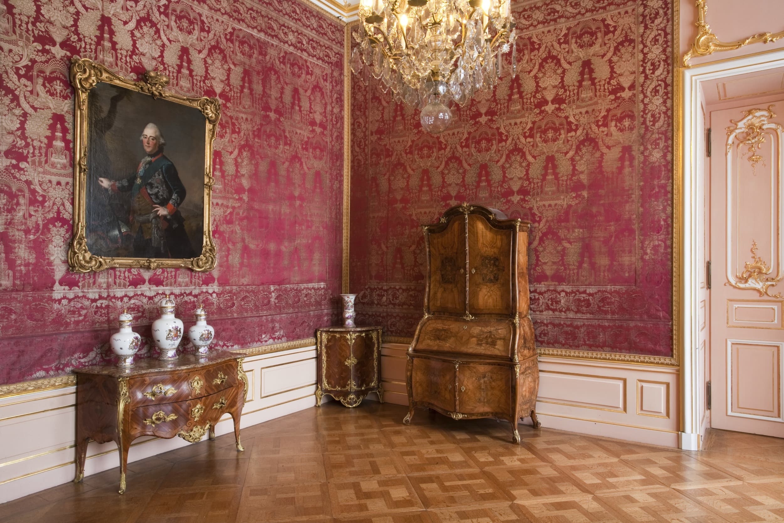 Verzierter Raum mit rot gemusterten Wänden, einem Kronleuchter, antiken Möbeln, Vasen und einem Porträt in einem kunstvollen Rahmen.