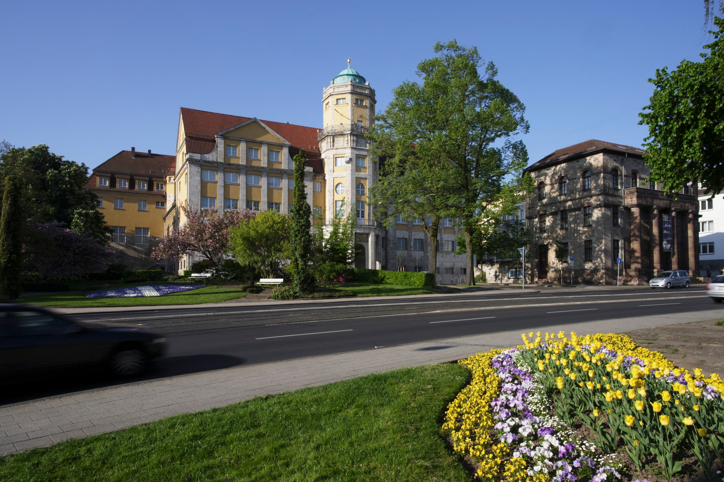 Historisches Gebäude mit Turm und rotem Dach, umgeben von Bäumen und bunten Blumen, neben einer Straße mit einem fahrenden Auto.