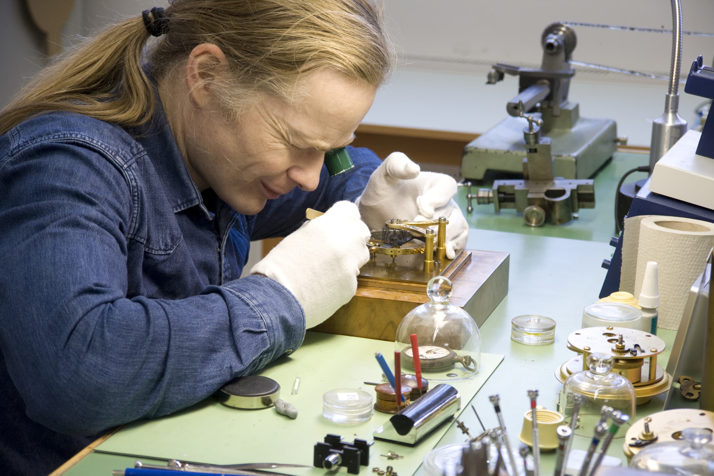 Eine Person trägt Handschuhe und arbeitet mit einer Lupe an einem kleinen mechanischen Gerät an einer überfüllten Werkbank.