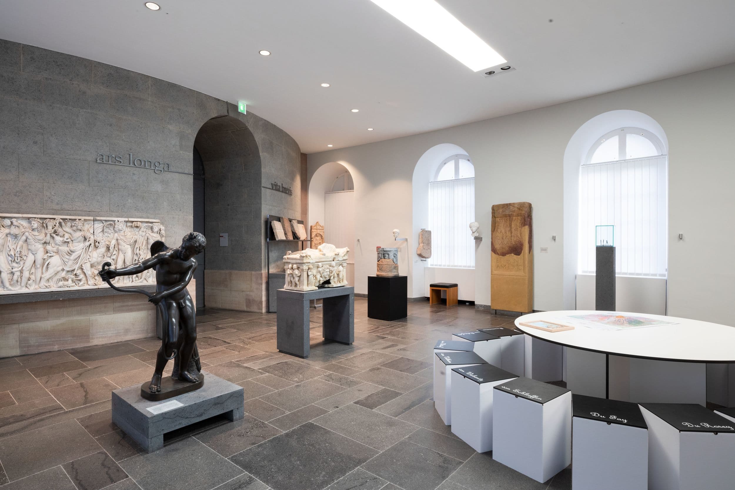 Eine Museumsgalerie mit Skulpturen, Wandkunst und Informationstafeln. Durch Bogenfenster fällt natürliches Licht ein und beleuchtet die Exponate.