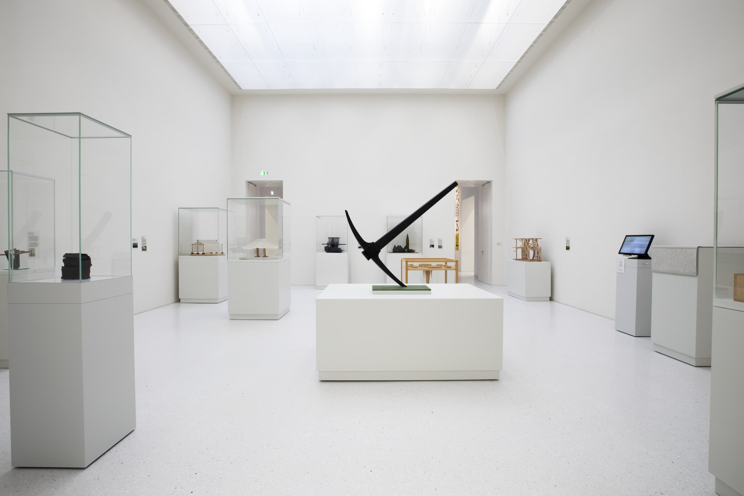 Minimalistische Kunstgalerie mit Skulpturen in Glasvitrinen, einem zentralen schwarzen abstrakten Stück auf einem weißen Sockel und einer hellen Deckenleuchte.