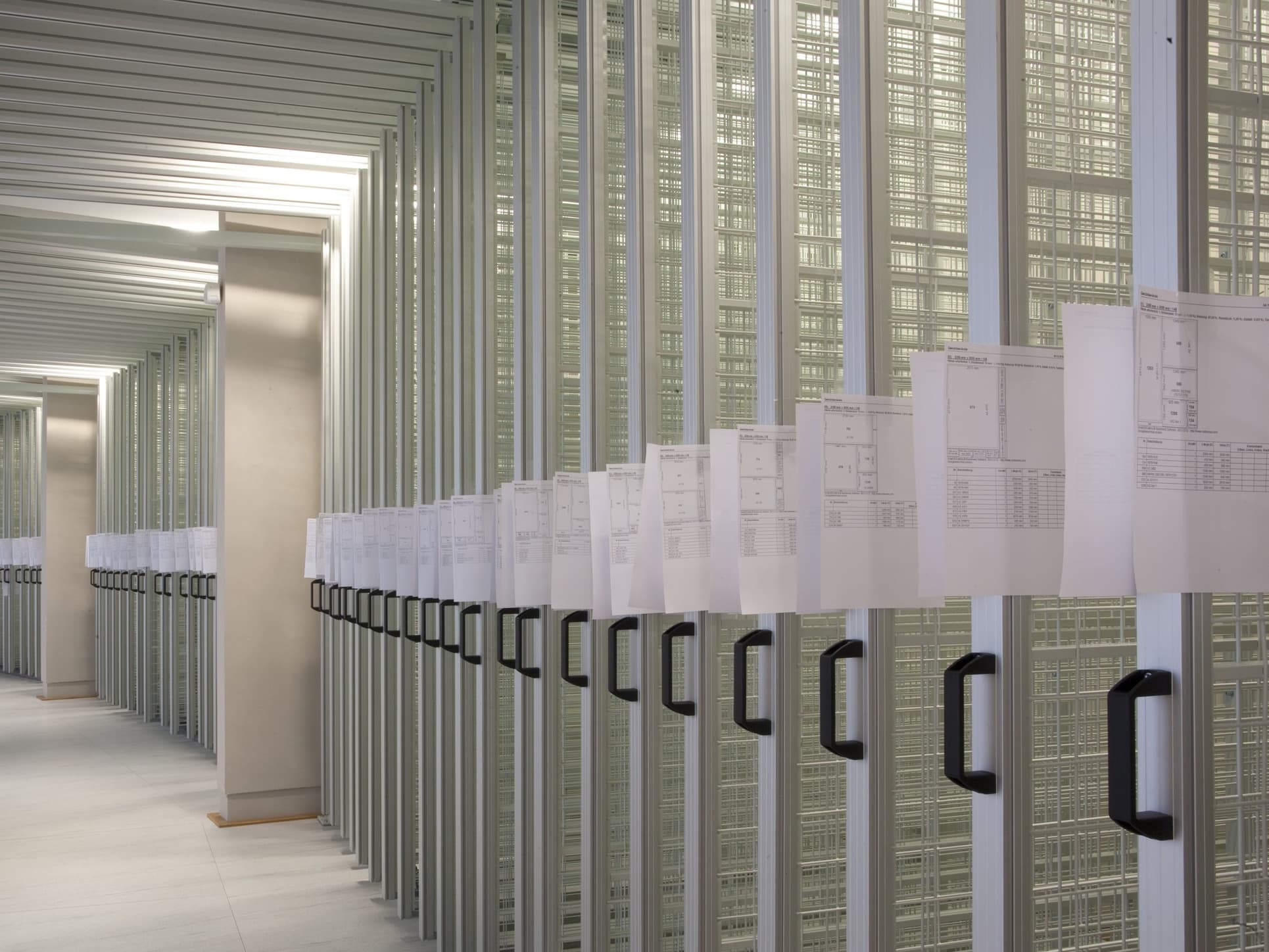 Reihen aus verschiebbaren Metallregalen mit Griffen in einem modernen Archiv oder einer Bibliothek, auf denen Dokumente sichtbar sind.