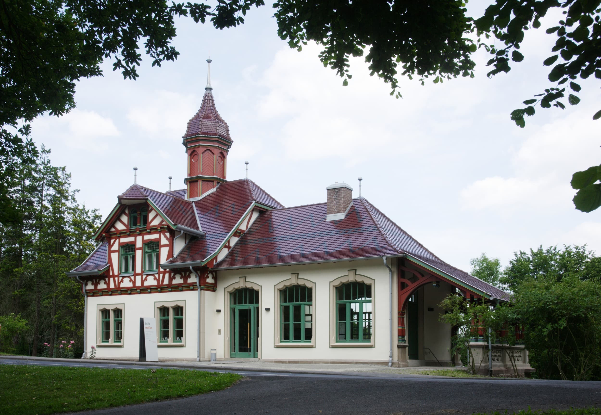 Ein malerisches, historisches Gebäude mit rotem Dach und verziertem Turm, umgeben von Grün unter einem teilweise bewölkten Himmel.