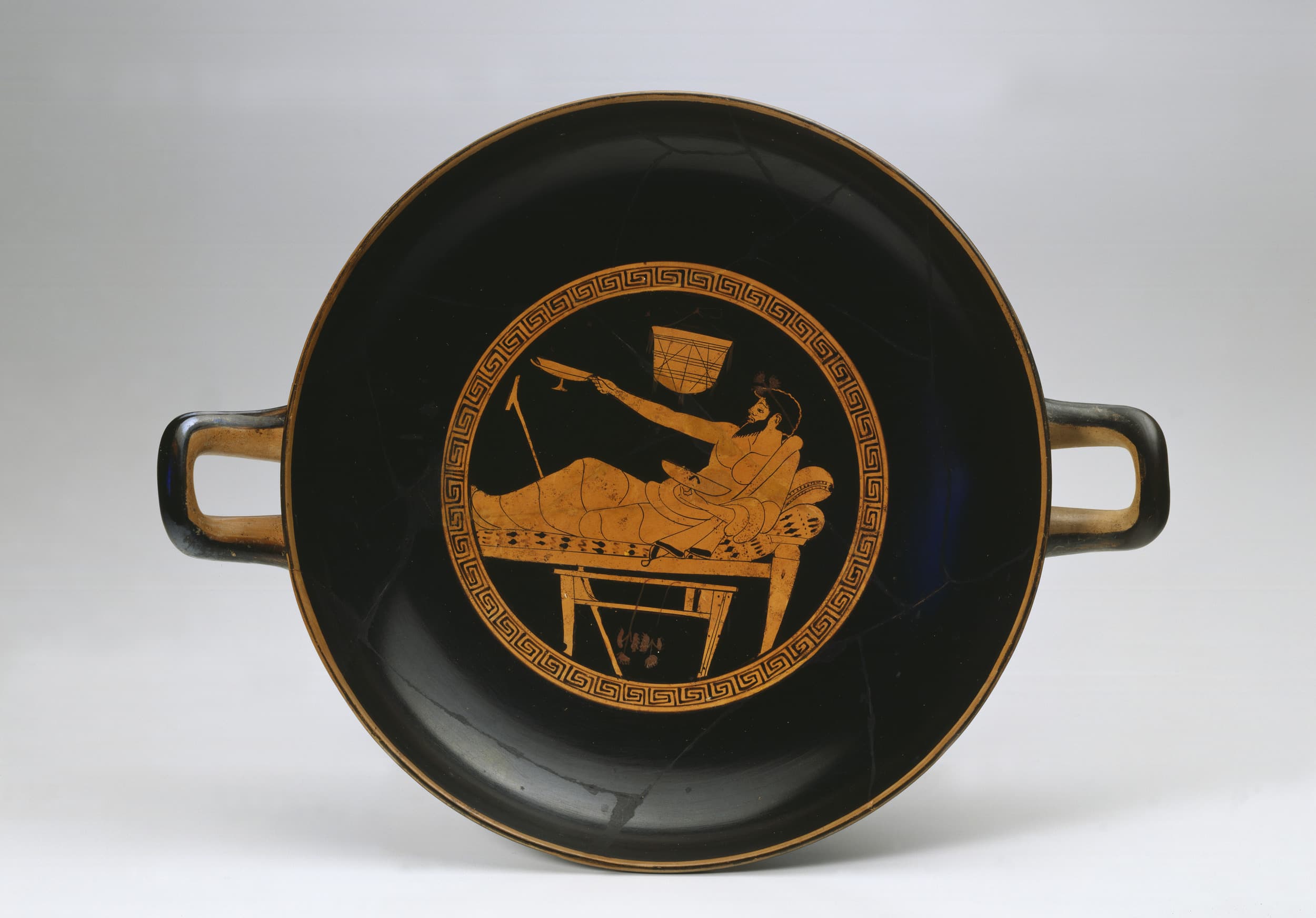 Antike griechische Kylix mit einer liegenden Figur, die eine Tasse hält, umgeben von einem geometrischen Rand und mit zwei Henkeln auf beiden Seiten.