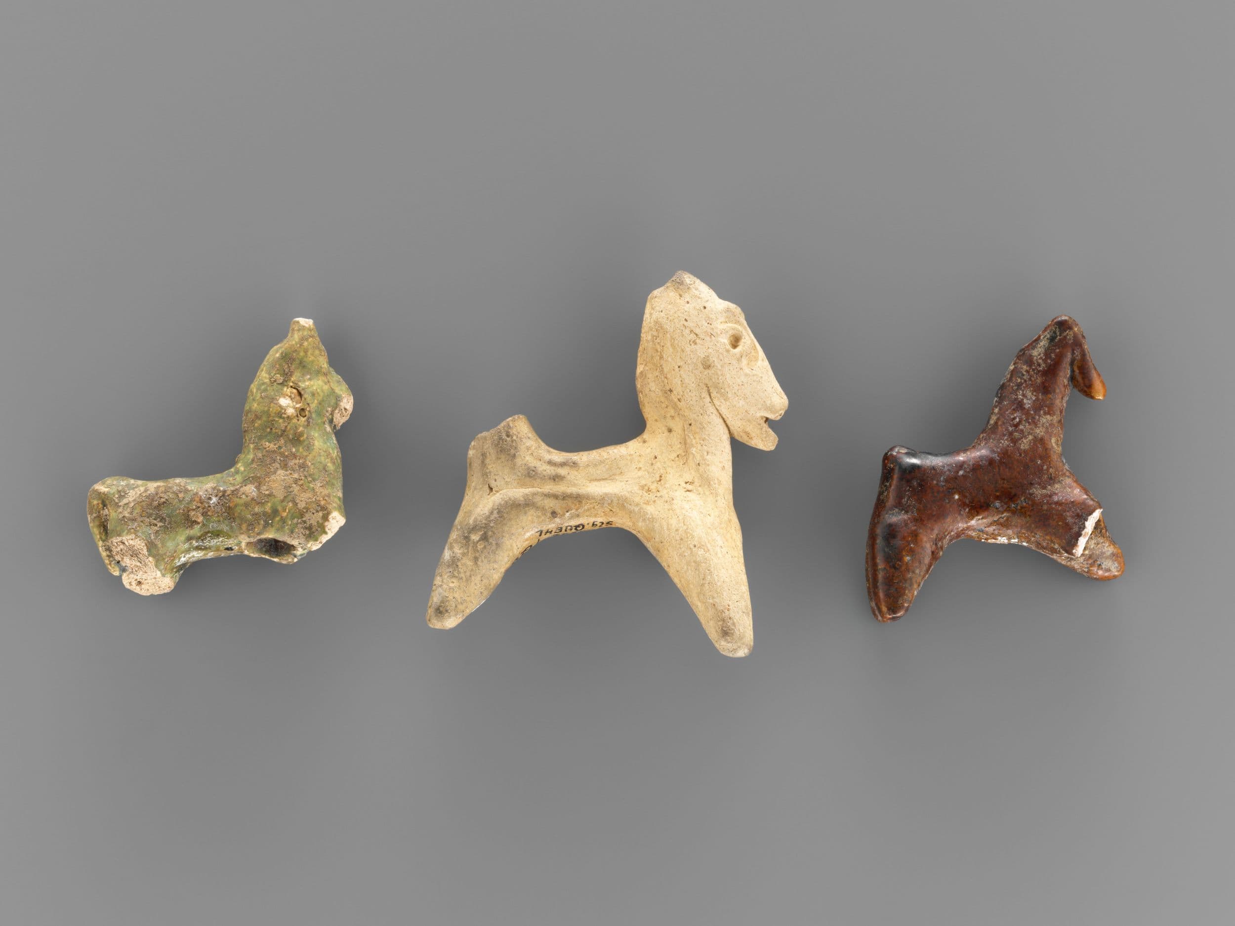 Three small, ancient horse figurines in varying shades of brown and beige, displayed on a plain gray background.