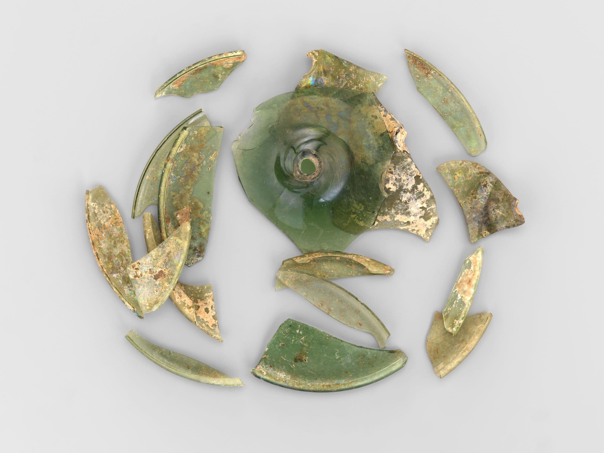 Broken pieces of at ancient green glass vessel arranged in a circular pattern on a gray background.