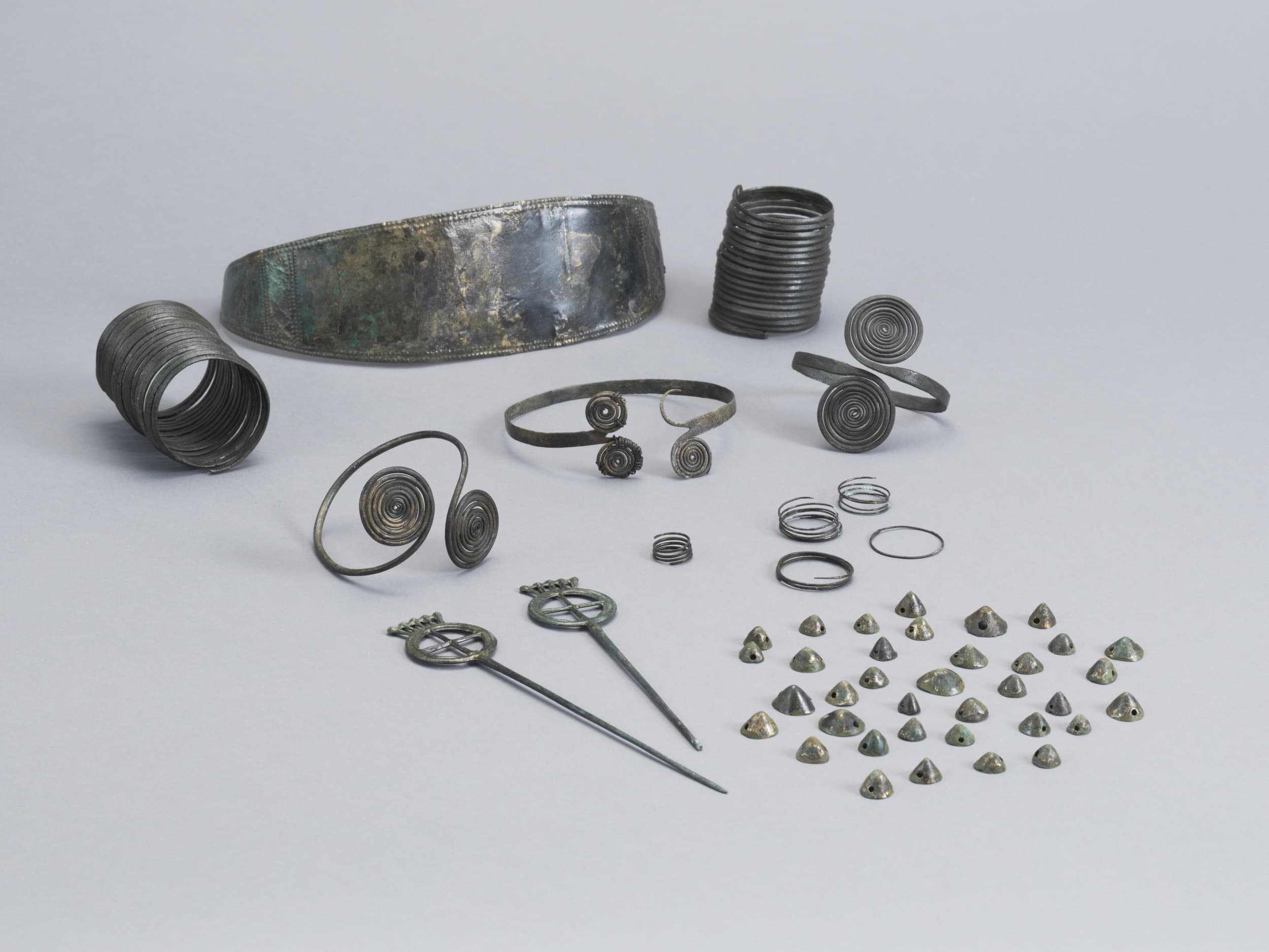 Eine Sammlung antiken Metallschmucks, darunter Armbänder, Ringe, Spiralmuster, Haarnadeln und kleine pyramidenförmige Stücke auf weißem Hintergrund.