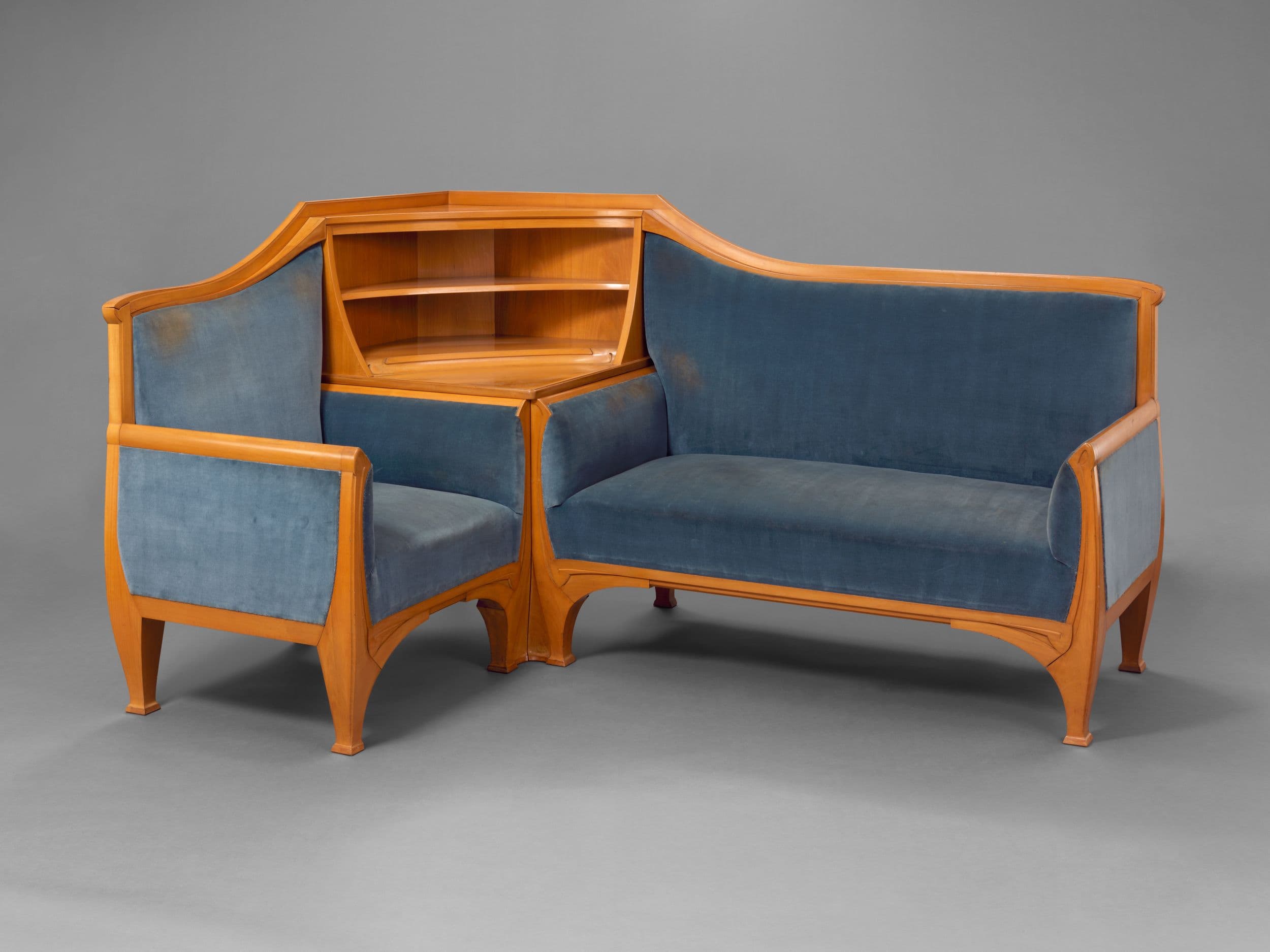 Ecksofa aus Holz mit blauer Polsterung und integrierten Regalen, bestehend aus einem Sessel und einem Zweisitzer-Element vor einem grauen Hintergrund.