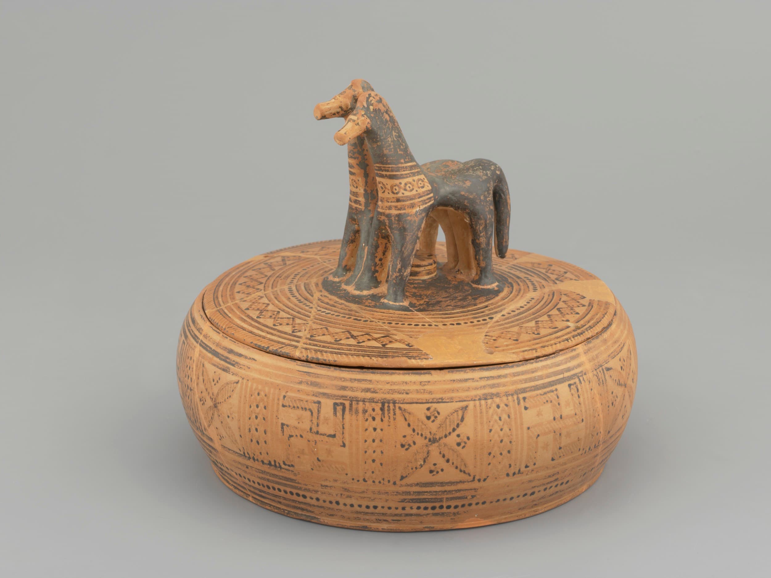 Ein runder, aufwendig gemusterter Terrakottabehälter mit einem Deckel, auf dem sich eine Pferdefigur befindet.