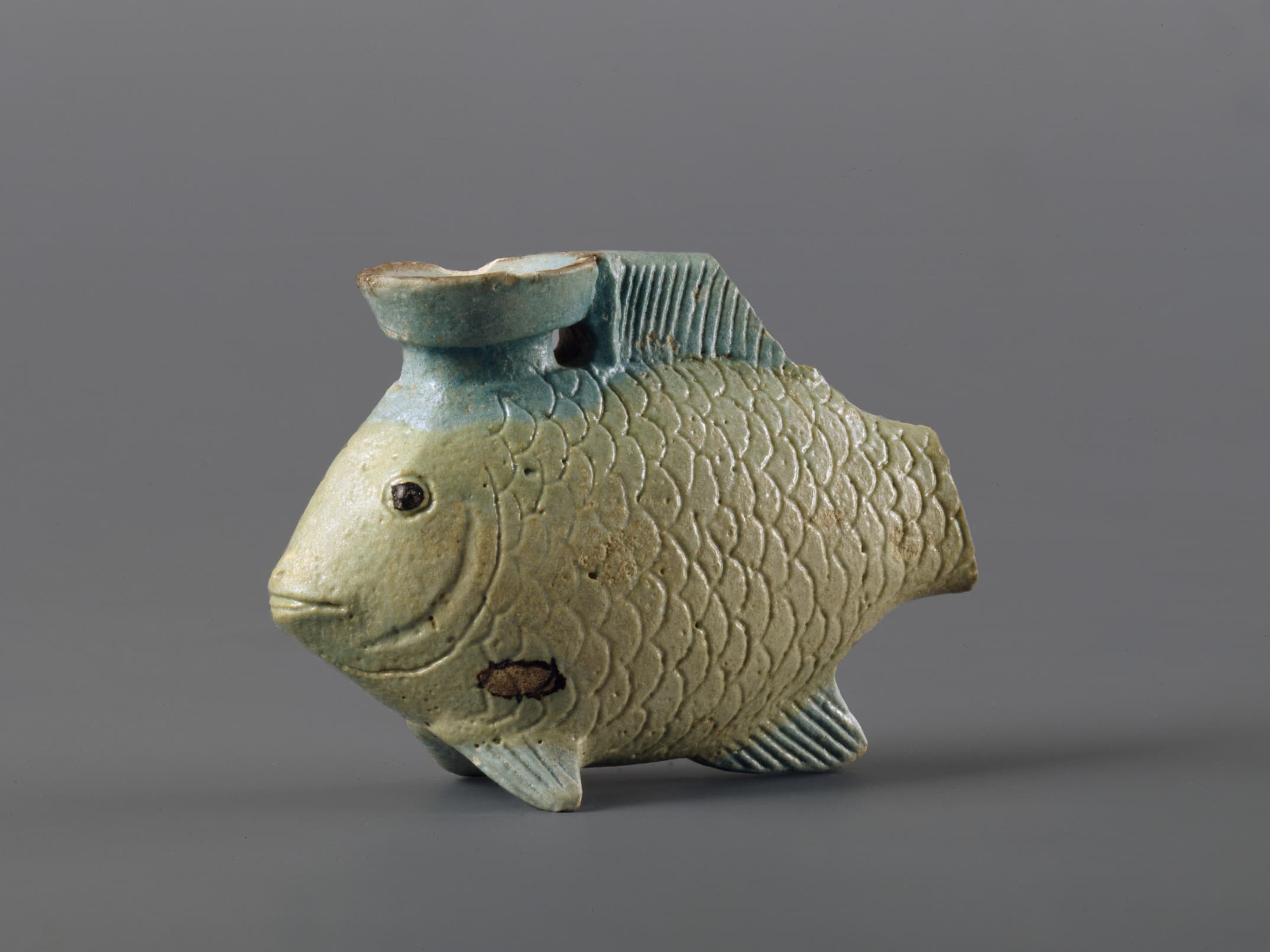 Fischförmiges Keramikgefäß mit detaillierten Schuppen, einer kleinen Öffnung oben und einer hellblaugrauen Glasur vor einem schlichten Hintergrund.