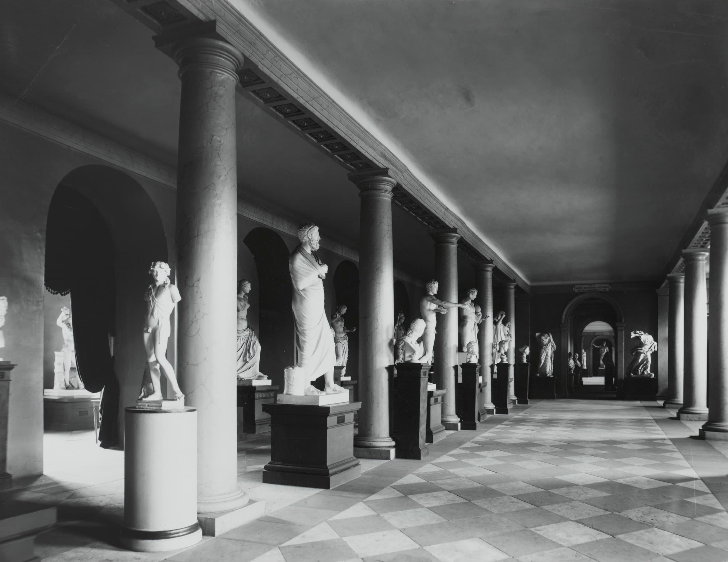 Ein Schwarzweißfoto einer Galerie mit klassischen Statuen auf Sockeln, flankiert von Säulen und einem karierten Boden.