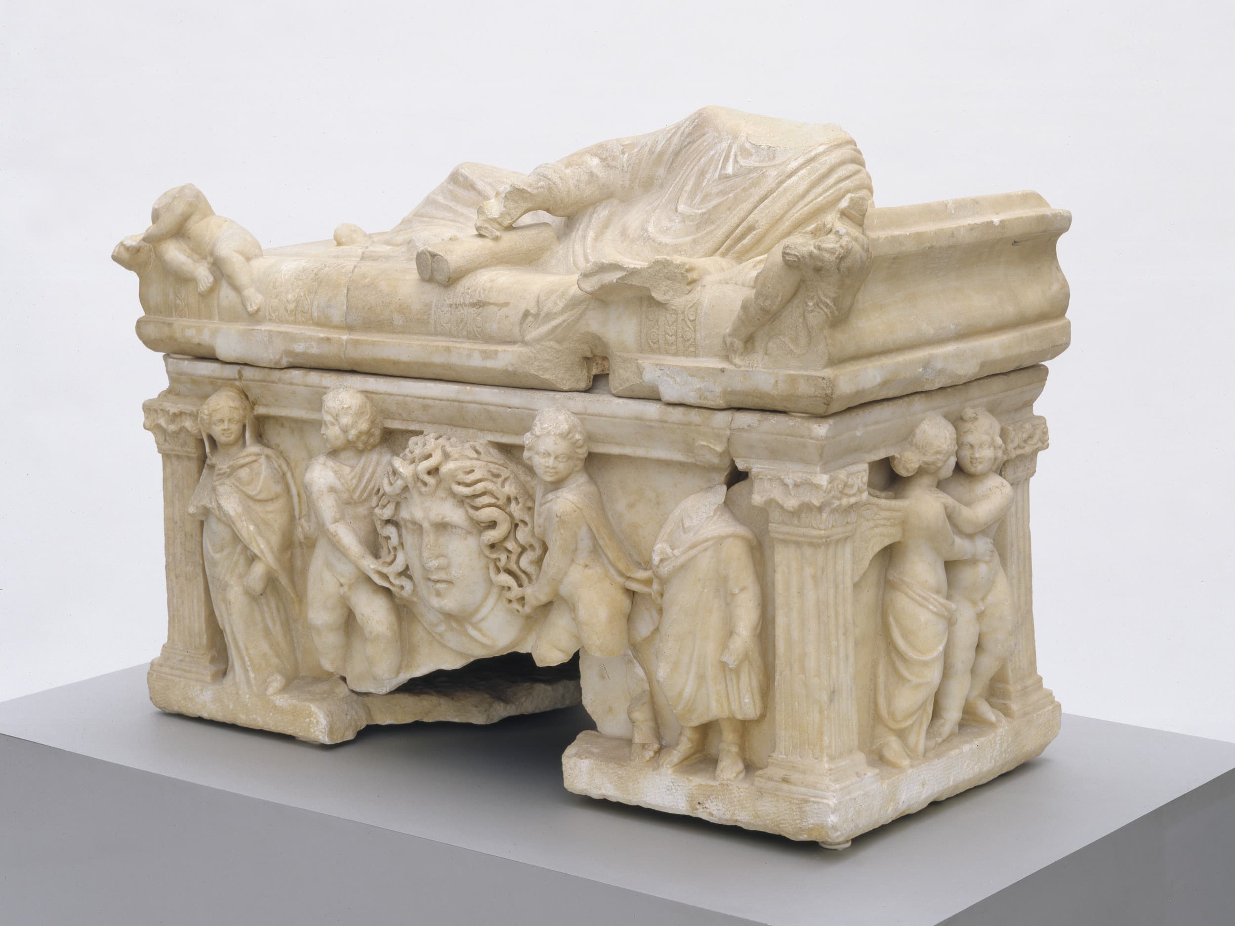 Antiker Marmorsarkophag mit liegender Figur auf der Oberseite, verziert mit Reliefs von Figuren und einem zentralen Medusenkopf, ausgestellt auf einem Sockel.