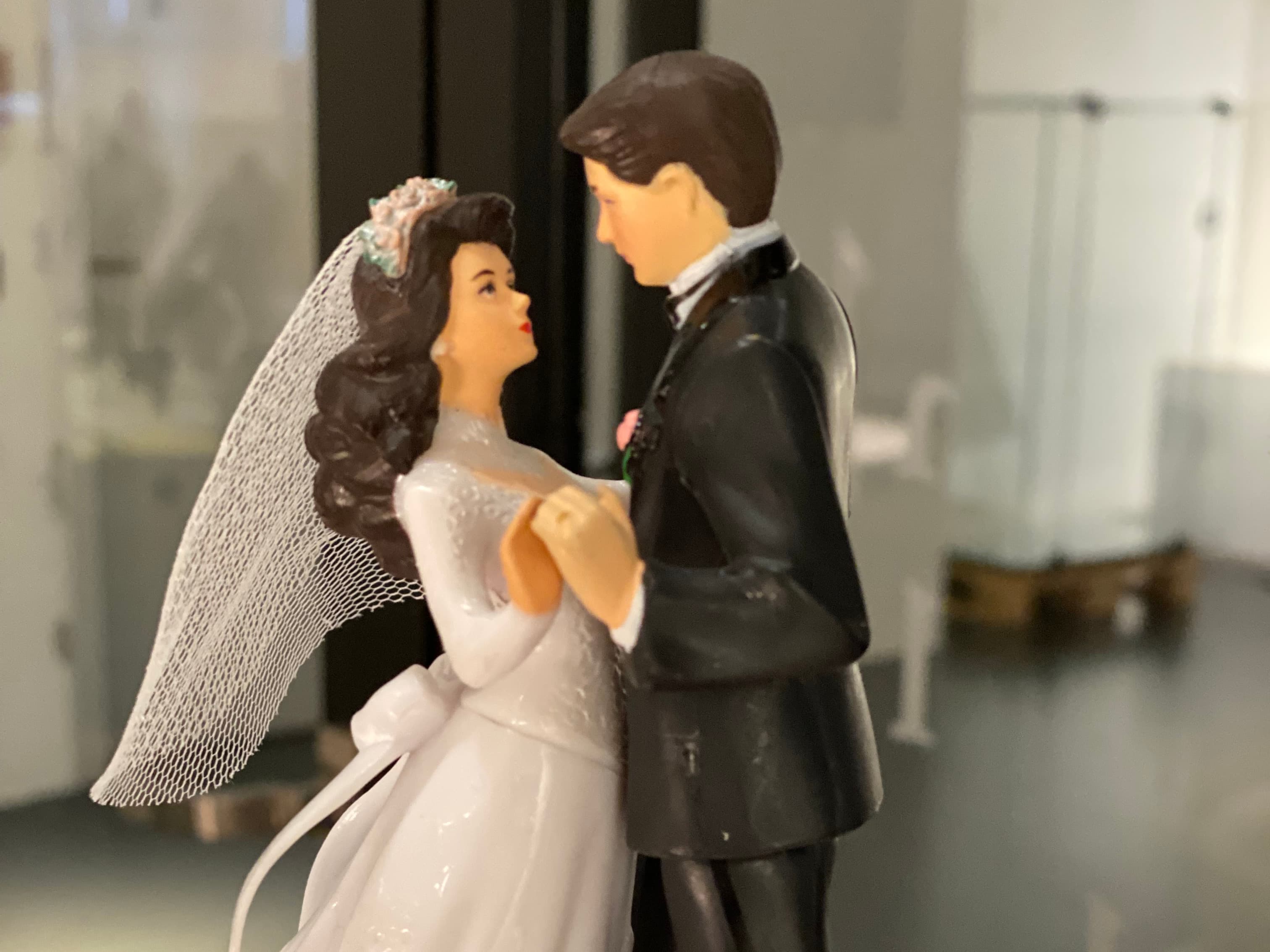 A bride and groom figurine in wedding attire, holding hands and gazing at each other, with a blurred background.