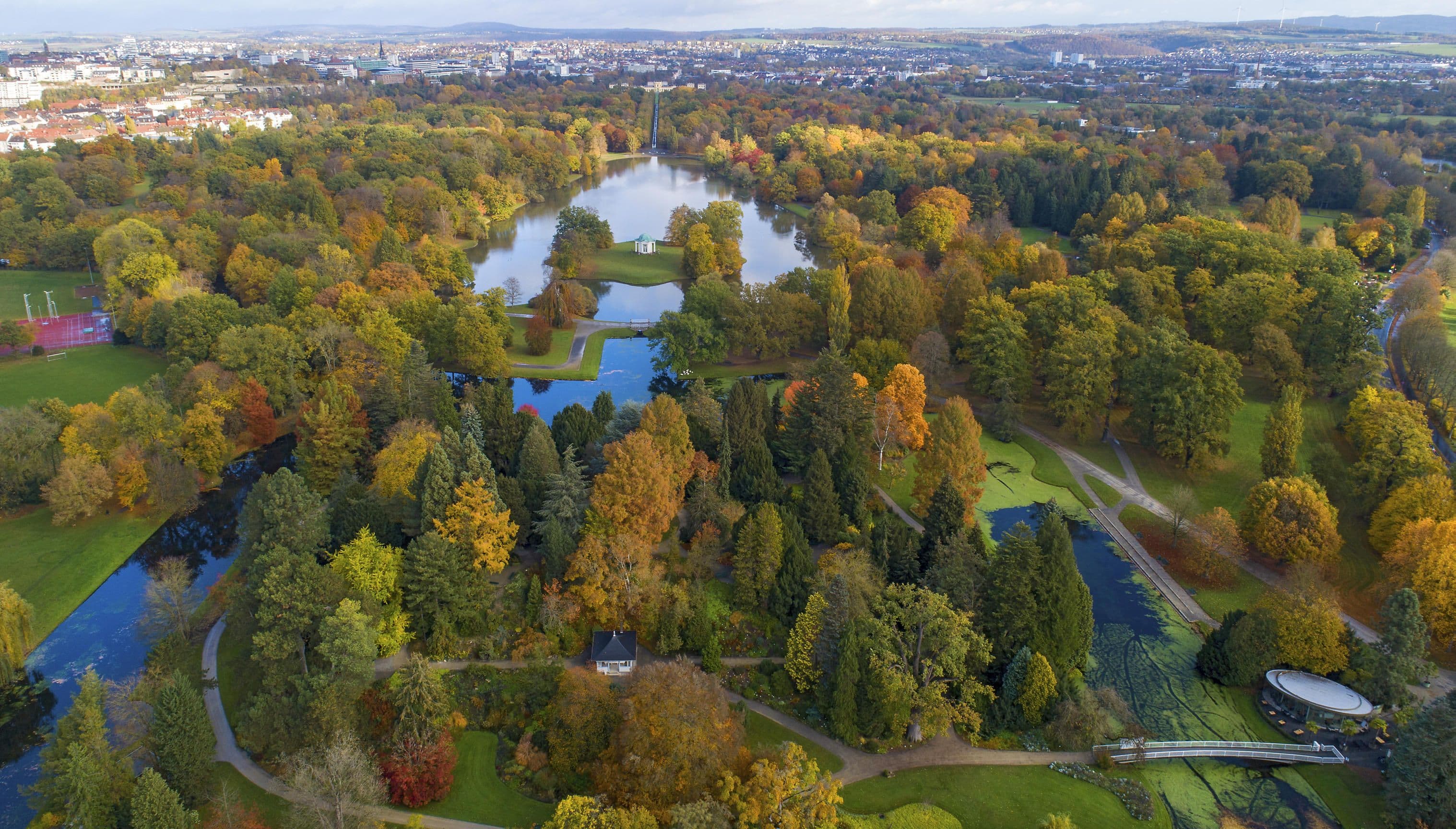 Aerial view of a lush park with vibrant autumn foliage, winding paths, and small lakes, set against a cityscape in the background.