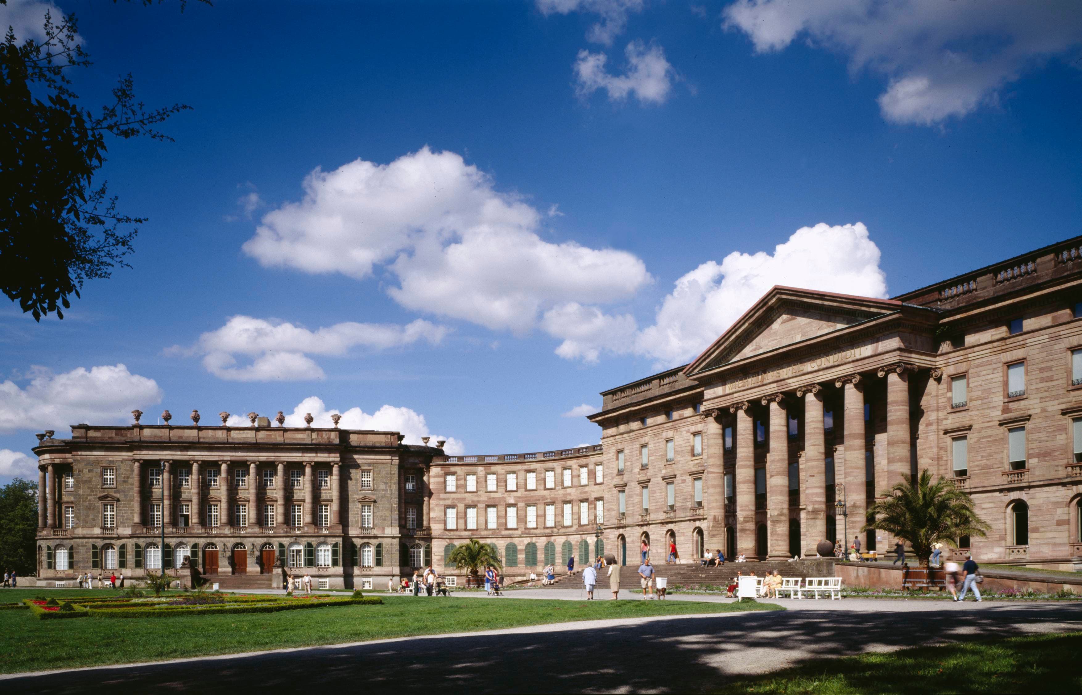 Historisches neoklassizistisches Gebäude mit großen Säulen, umgeben von einem grünen Rasen und Menschen, die unter einem blauen Himmel mit vereinzelten Wolken spazieren gehen.