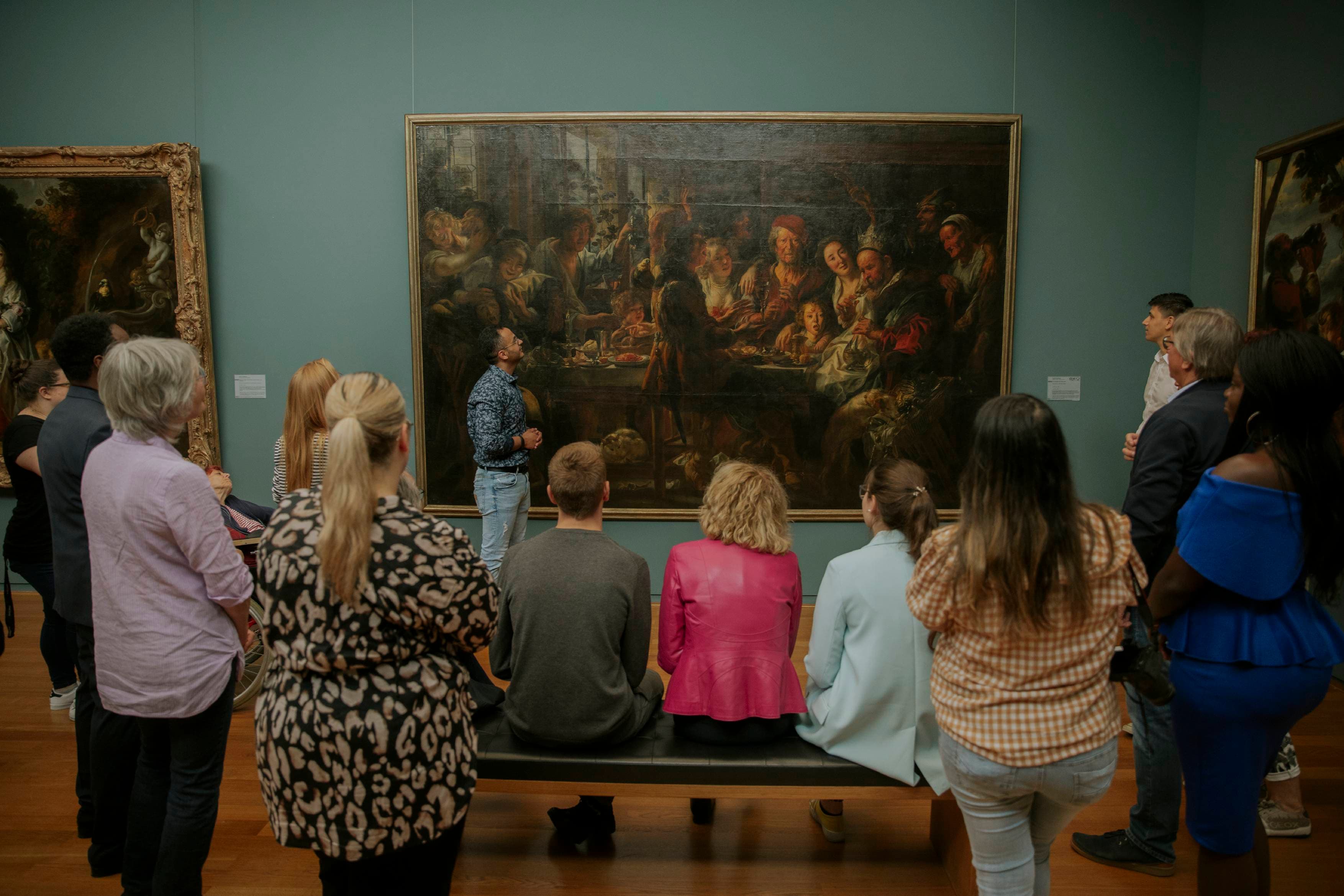 Eine Gruppe von Menschen betrachtet ein großes Gemälde in einer Museumsgalerie, während ein Führer das Kunstwerk erklärt.