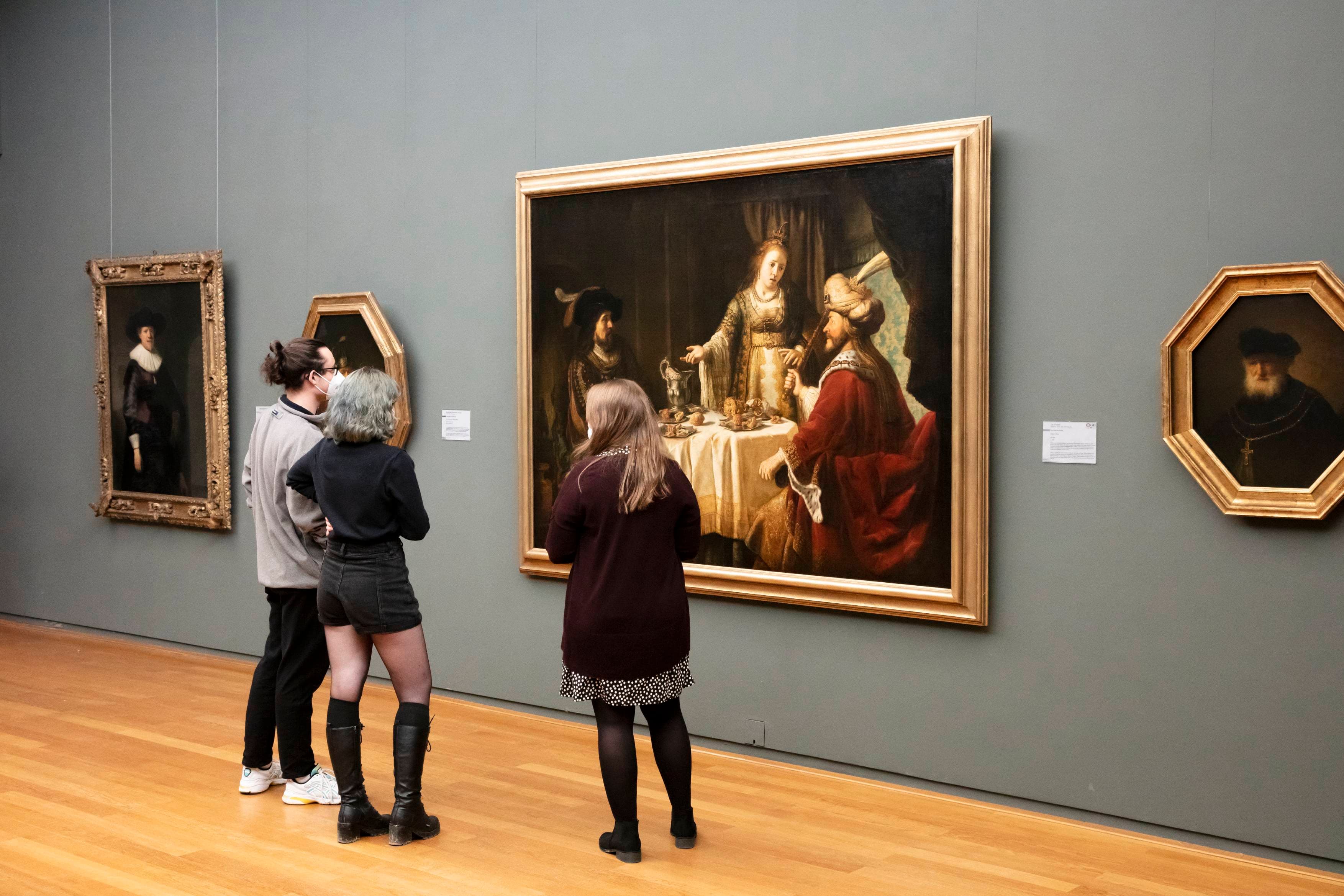 Drei Personen betrachten gerahmte Gemälde in einer Kunstgalerie und konzentrieren sich auf ein großes, detailliertes Stück, das eine historische Szene darstellt.