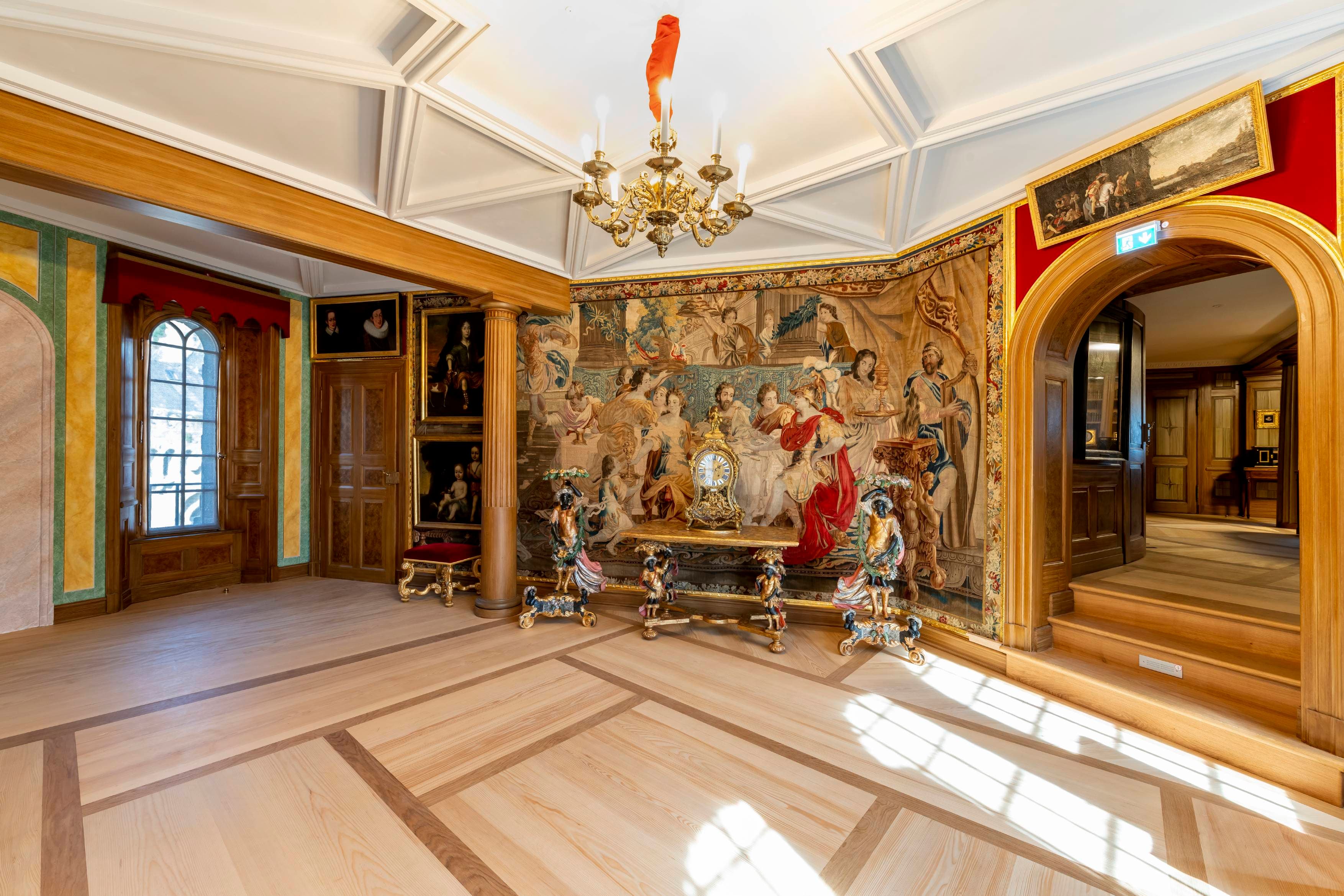 Reich verzierter Raum mit großem Wandteppich, Kronleuchter, gewölbter Tür und Holzboden. Sonnenlicht fällt durch ein hohes Fenster auf der linken Seite.