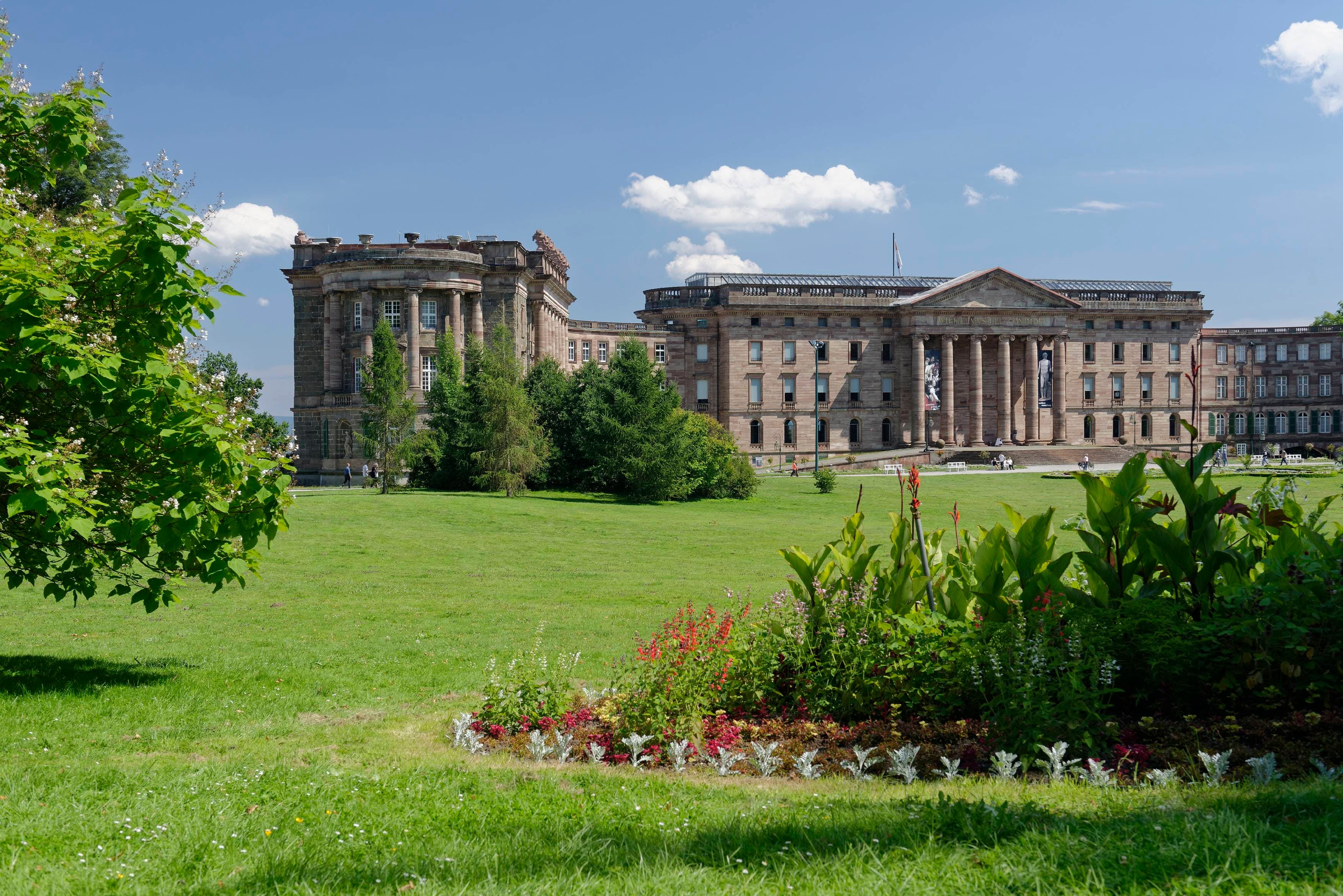 Historisches Gebäude mit Säulen und verzierter Fassade, umgeben von einem üppigen grünen Rasen und einem bunten Blumenbeet unter einem klaren blauen Himmel.
