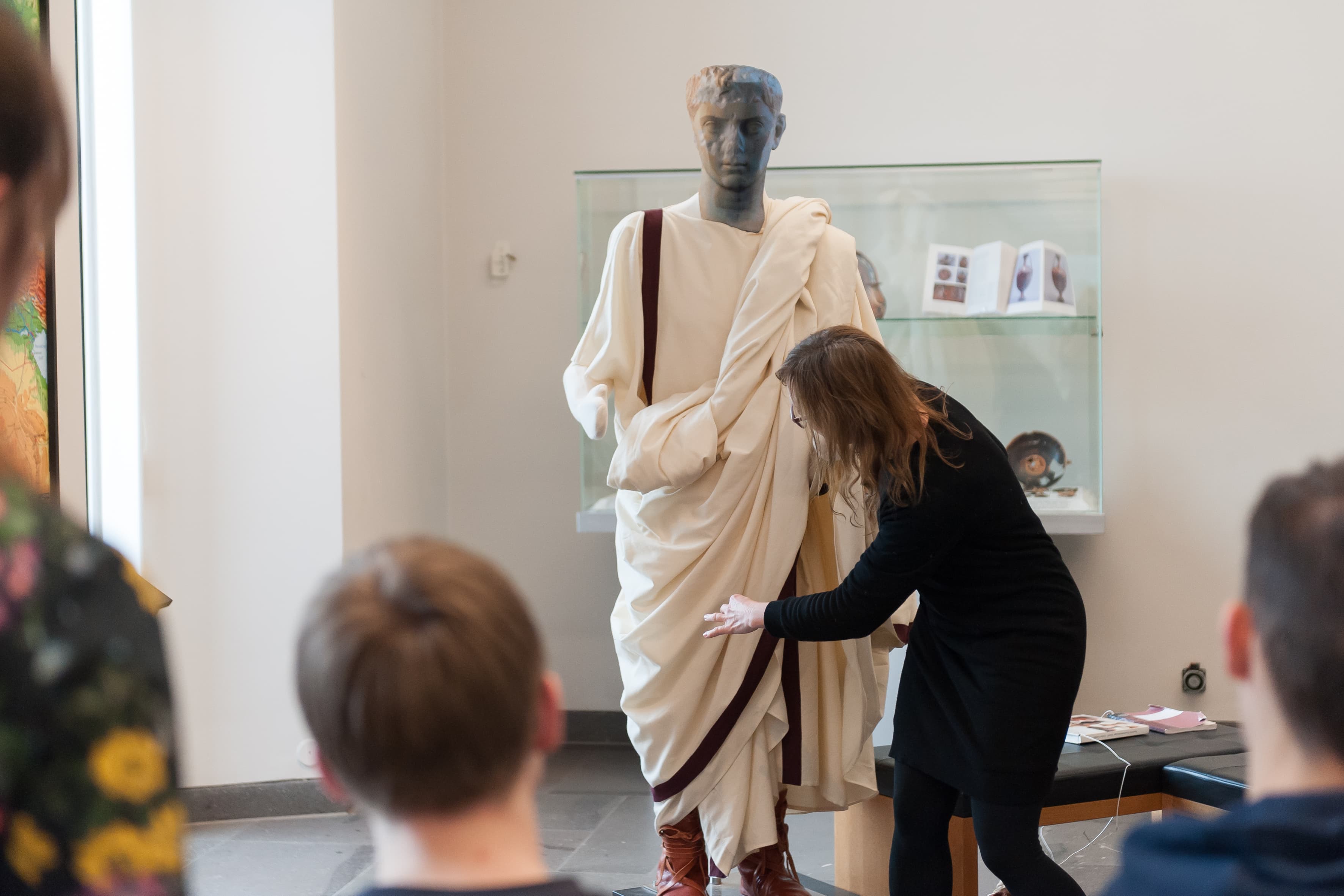 Führungskraft erklärt Schülerinnen und Schülern anhand einer Figurine das korrekte Tragen einer Toga