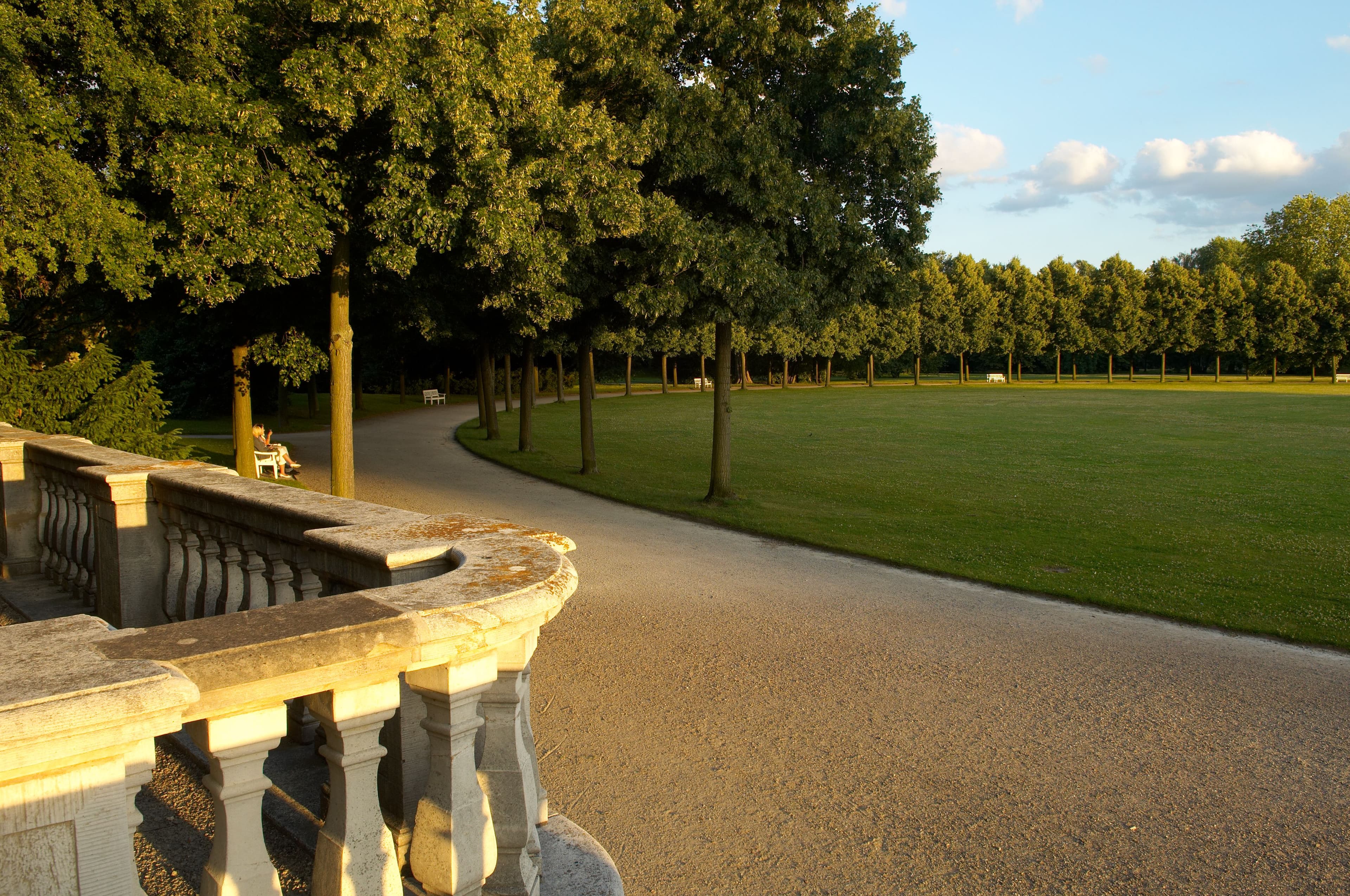A sunlit park scene with a curved gravel path, lined by trees, and a stone balustrade in the foreground.