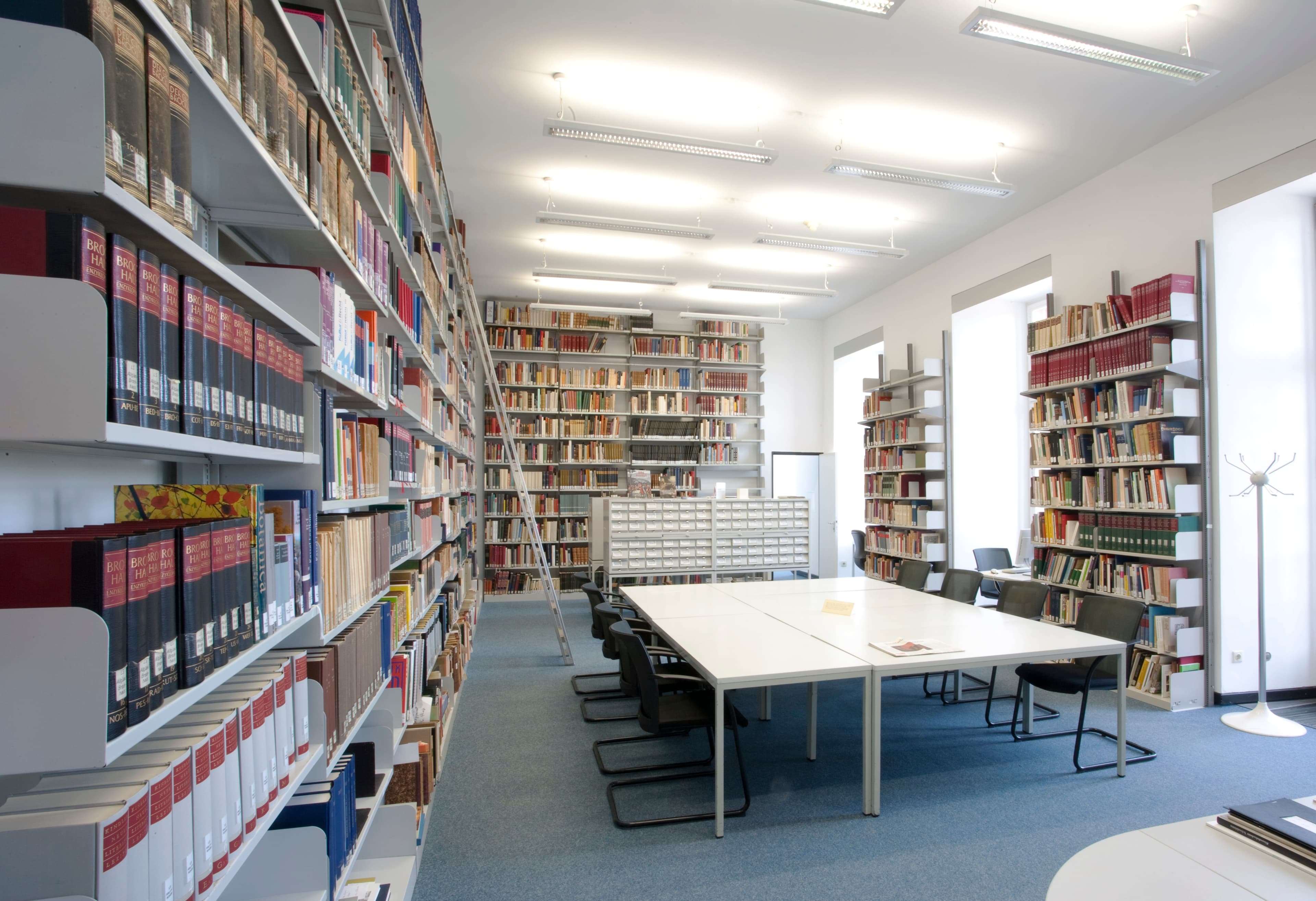 Ein heller Bibliotheksraum mit Bücherregalen, einem langen Tisch mit Stühlen und großen Fenstern, die natürliches Licht hereinlassen.