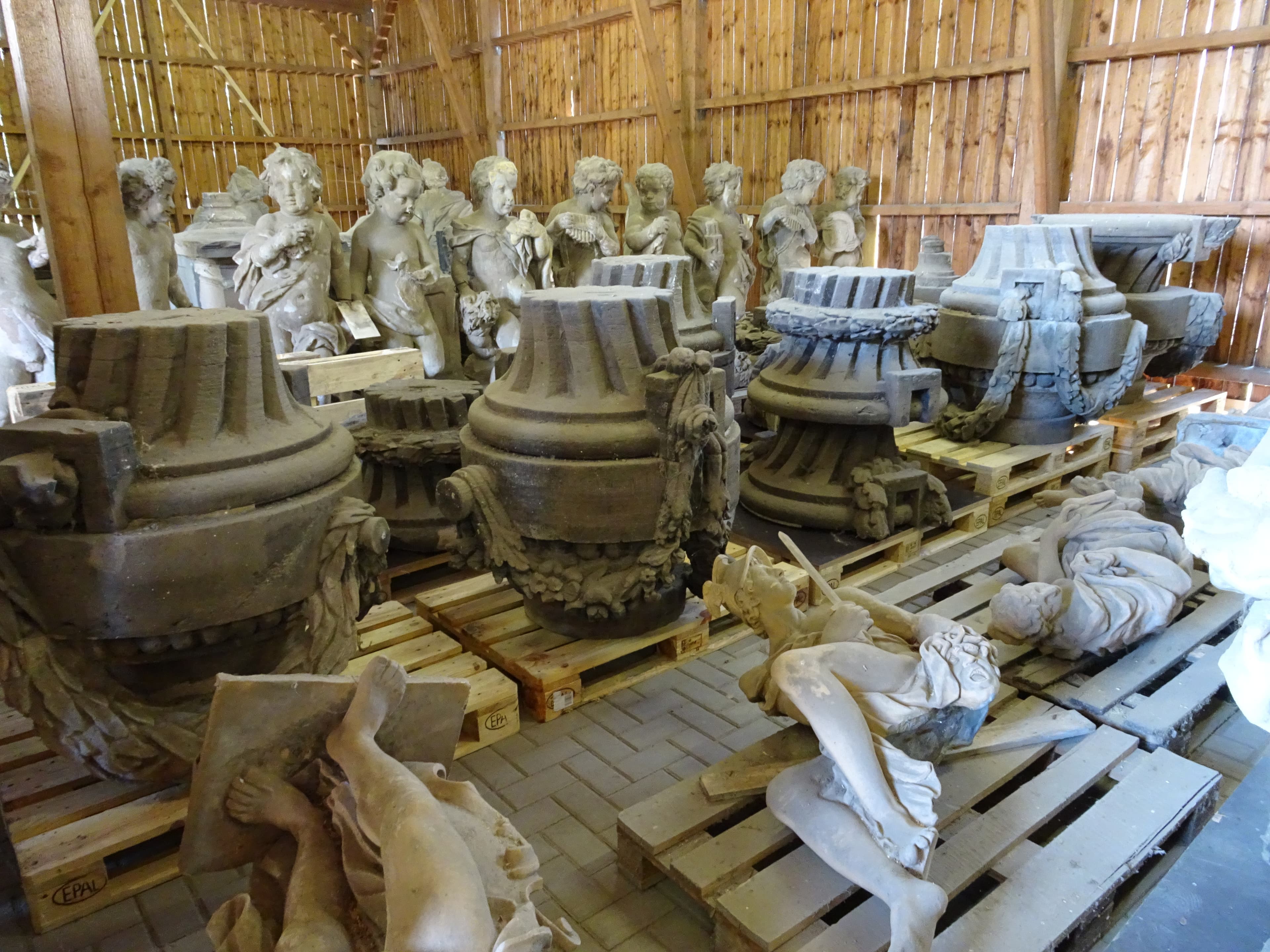 Steinskulpturen und große Urnen, die auf Holzpaletten in einem Holzlagerhaus gelagert sind. Mehrere Statuen und dekorative Elemente sind sichtbar.