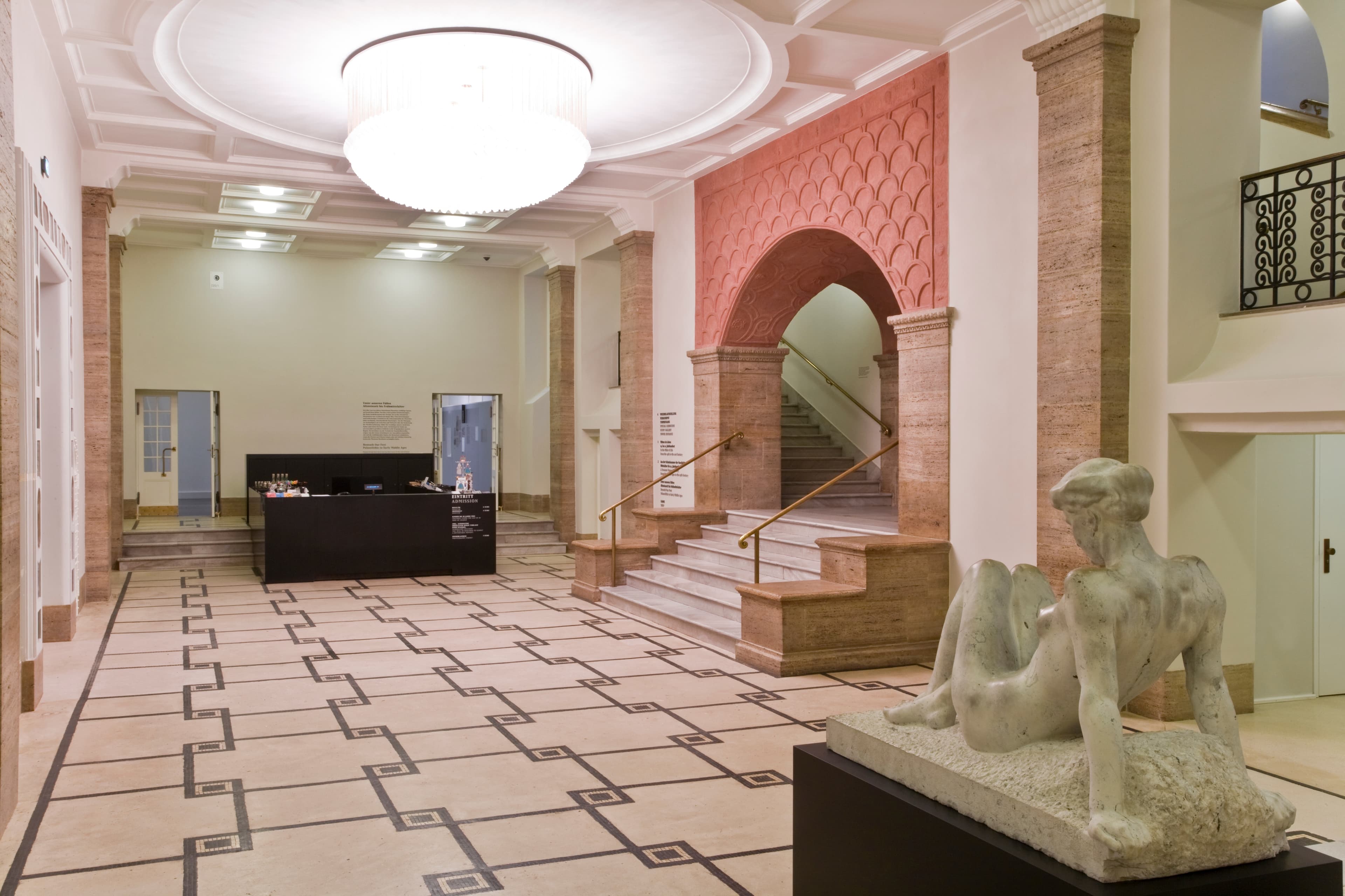 Geräumige Lobby mit modernem Kronleuchter, einer Skulptur auf einem Sockel und einer Treppe mit rotem Torbogen. Elegantes Dekor und Fliesenboden.