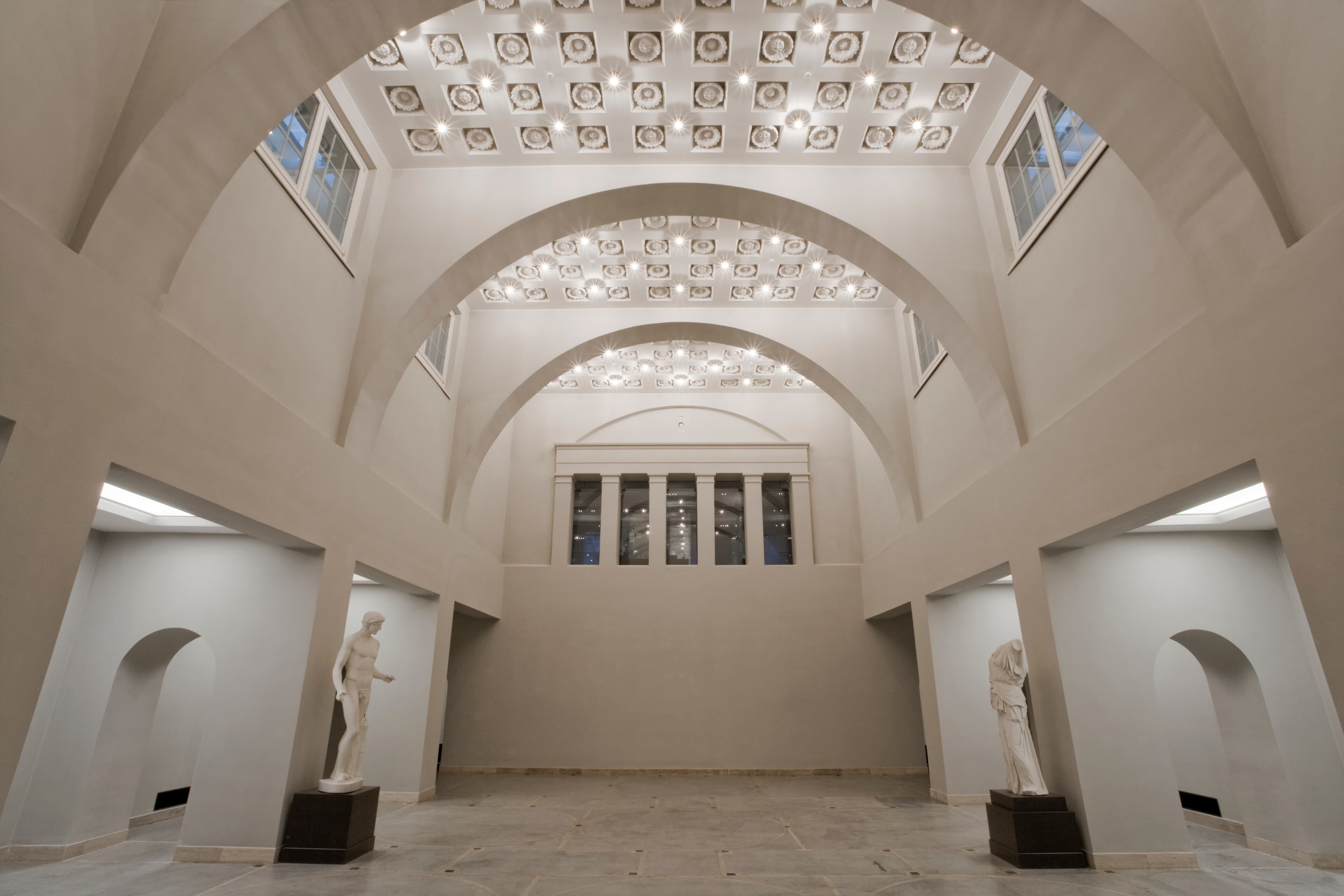 Geräumiger, eleganter Saal mit gewölbter Decke, dekorativer Beleuchtung und klassischen Statuen auf beiden Seiten. Neutrale Töne und minimalistisches Design.