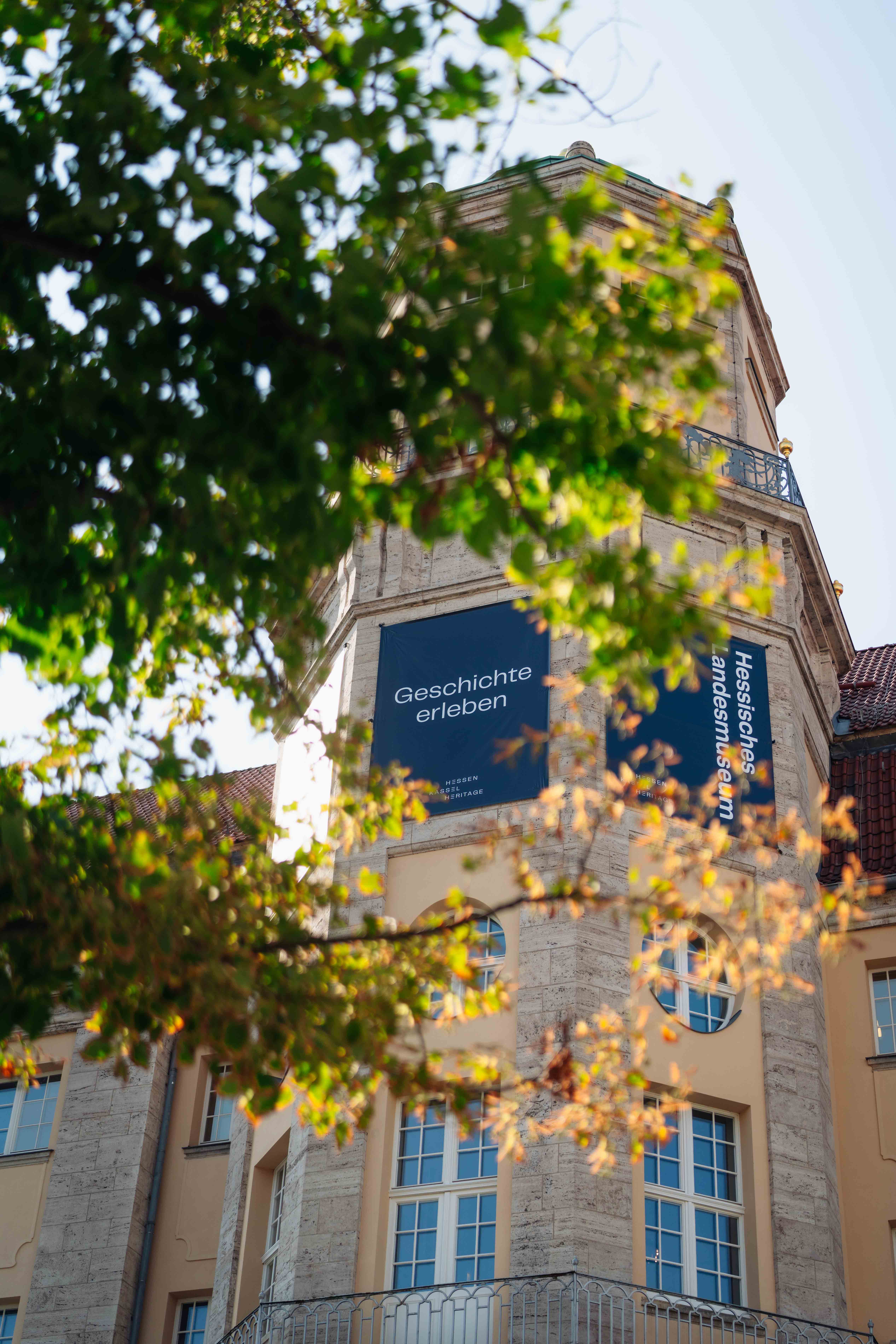 Historic building with a stone tower, partially obscured by tree branches. A sign on the tower reads "Geschichte erleben."