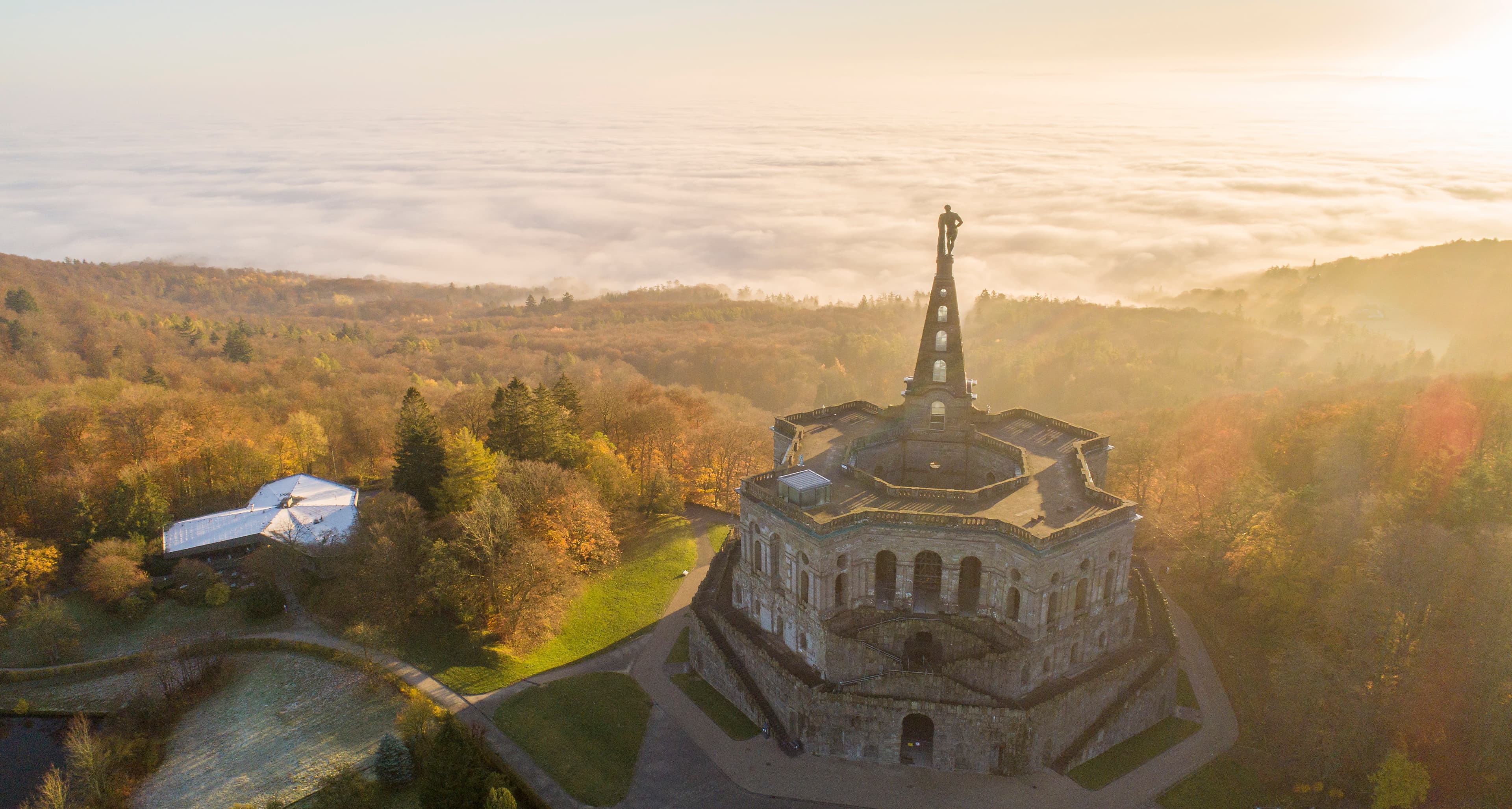 Luftaufnahme des Herkules-Denkmals in Kassel, Deutschland, umgeben von Herbstbäumen und einer nebligen Landschaft bei Sonnenaufgang.