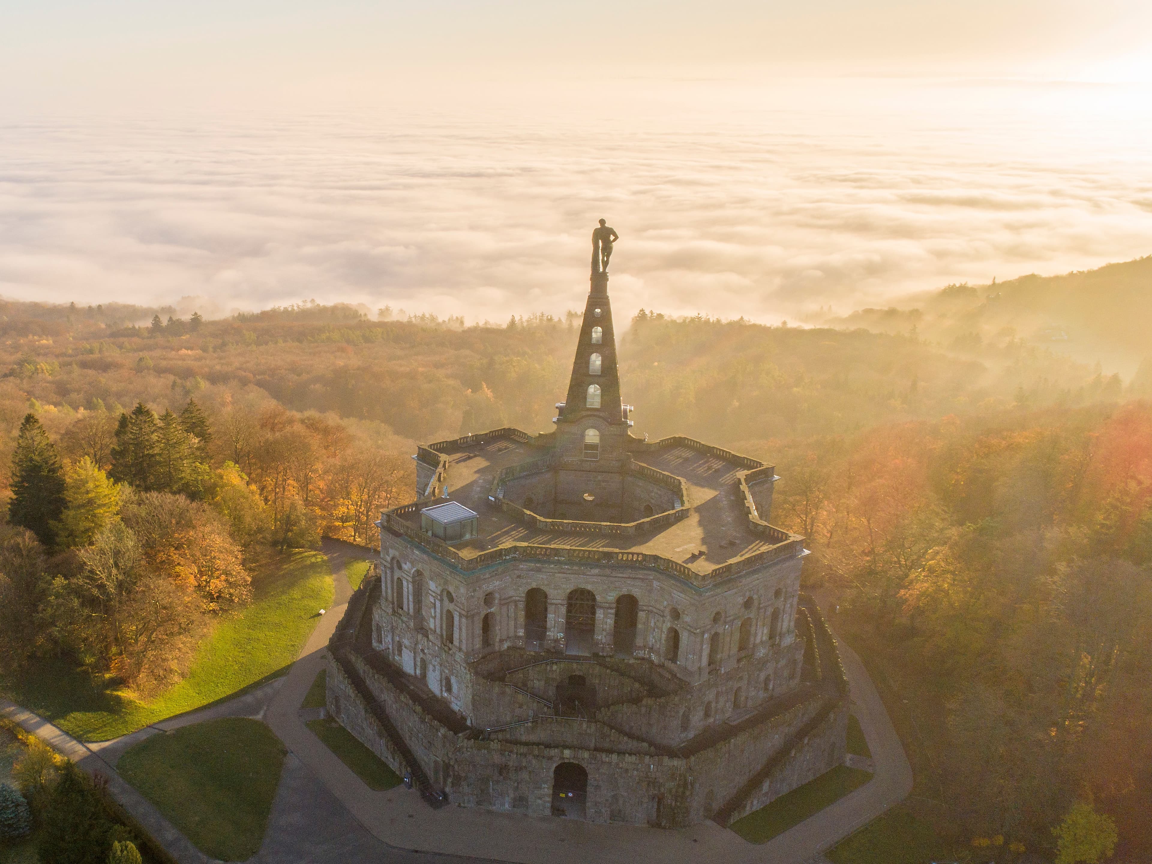 Luftaufnahme des Herkules-Denkmals in Kassel, Deutschland, umgeben von Herbstbäumen und einer nebligen Landschaft bei Sonnenaufgang.