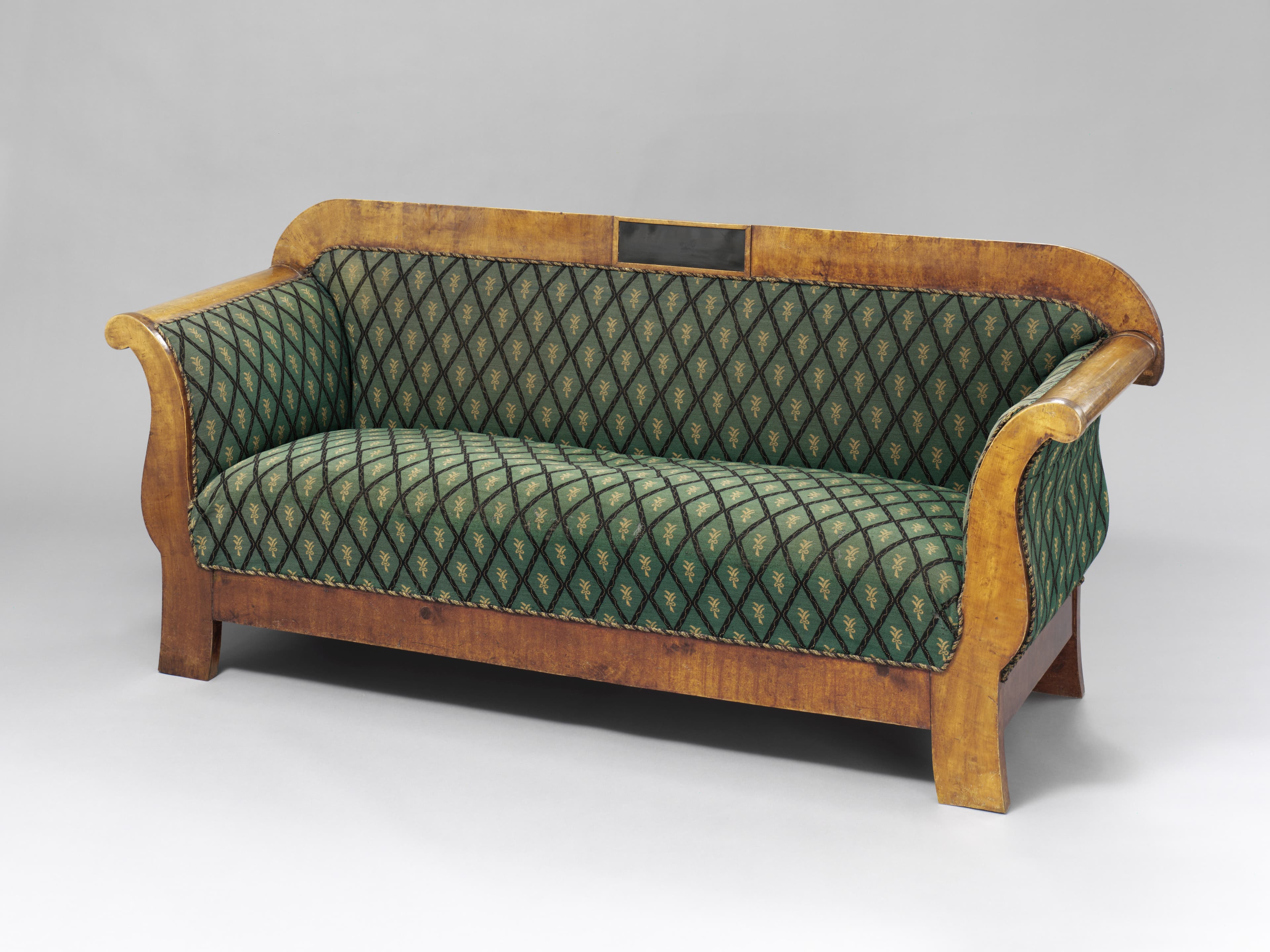 Antikes Holzsofa mit geschwungenen Armlehnen und grüner Polsterung mit Rautenmuster auf schlichtem grauem Hintergrund.