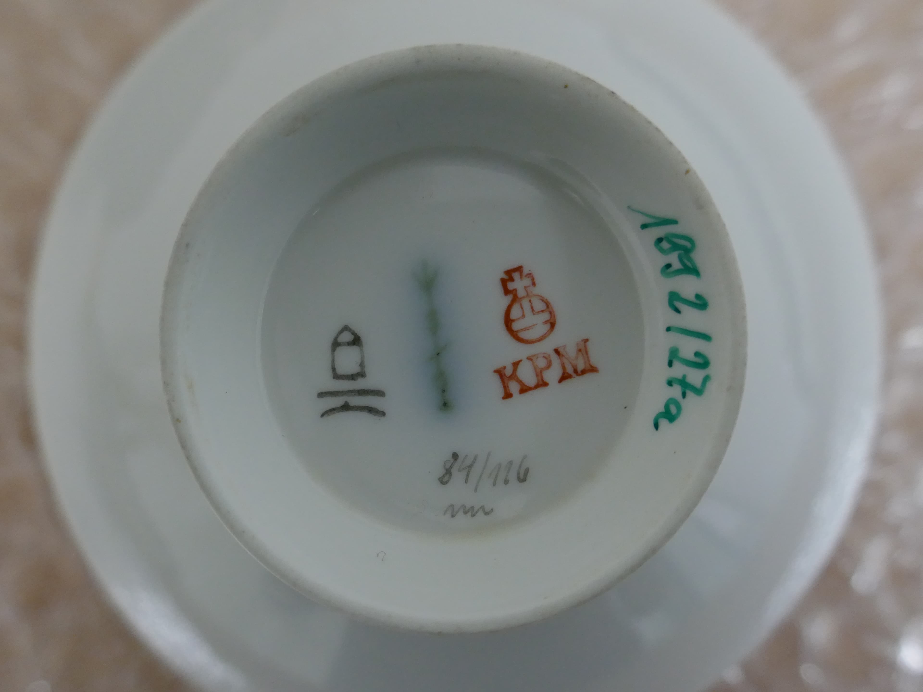 Sockel eines Porzellanartikels mit verschiedenen Markierungen, darunter ein rotes „KPM“-Logo, ein Haussymbol, Zahlen und eine grüne handschriftliche Notiz.