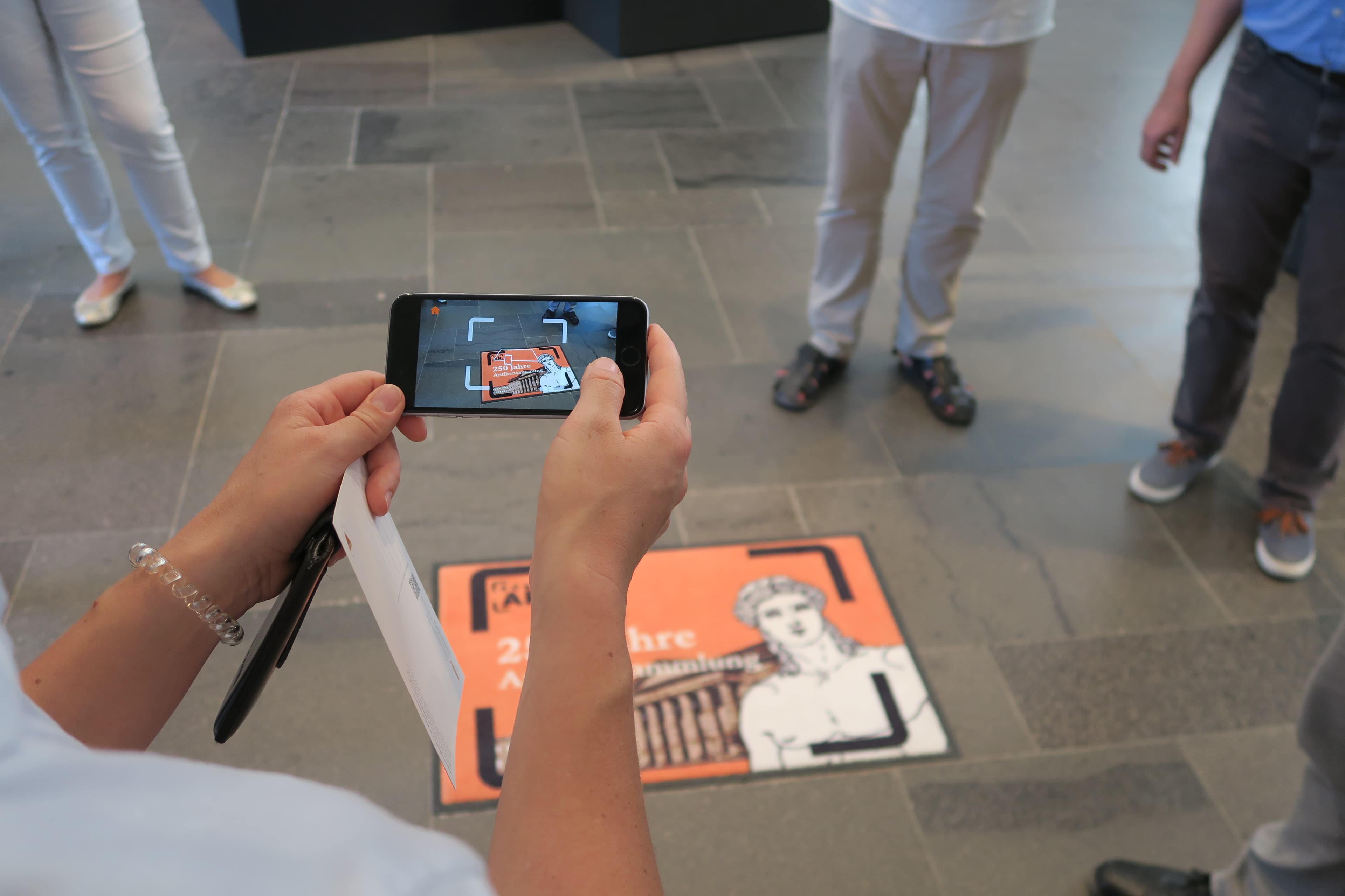 Eine Person hält ein Smartphone und scannt einen Bodenmarker in der Antikensammlung. Auf dem Smartphone-Display erscheint eine digitale Version des Markers mit dem Text '250 Jahre Antikensammlung'. Mehrere Menschen stehen um den Bodenmarker herum.