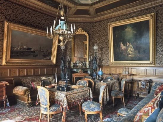 Verzierter Vintage-Raum mit gemusterter Tapete, großen Gemälden, Kronleuchter und antiken Möbeln, darunter ein Tischset mit Stühlen und Dekorationsgegenständen.