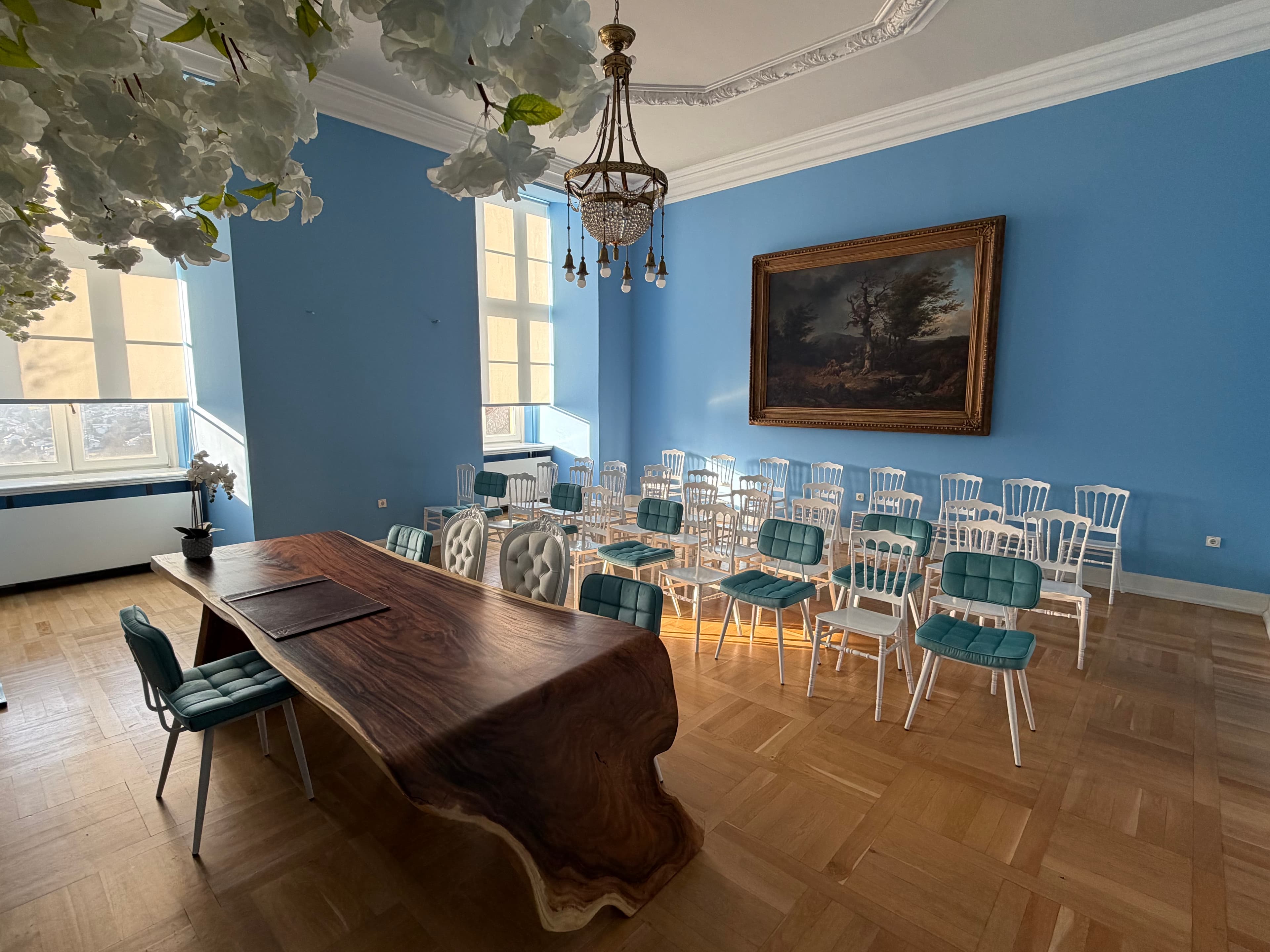 Elegantes Zimmer mit blauen Wänden, einem großen Holztisch, Stühlen in Reihen, einem Kronleuchter und einem Landschaftsgemälde. Helles Tageslicht strömt herein.