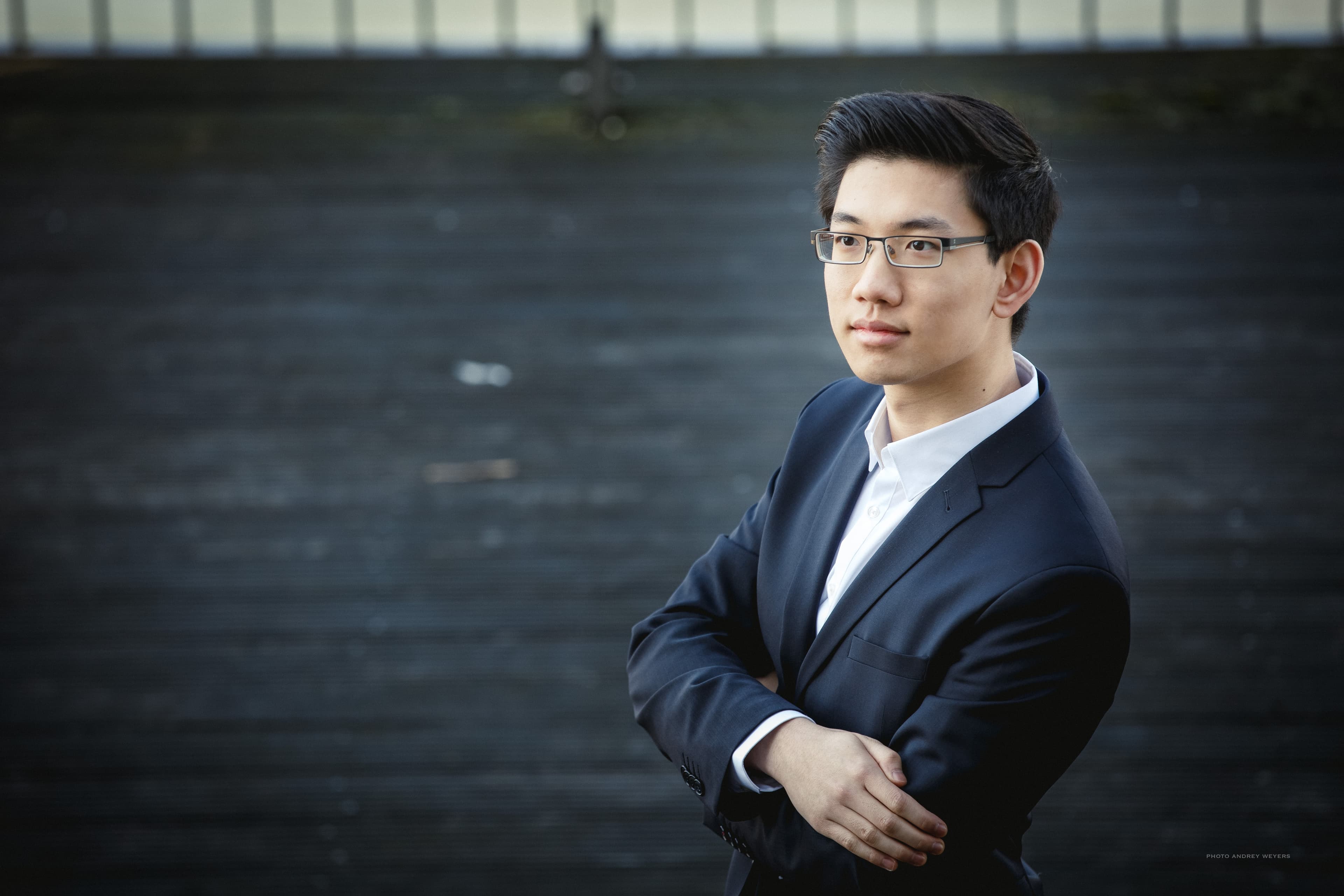 Ein Mann in Anzug und Brille steht selbstbewusst mit verschränkten Armen da und blickt vor einem dunklen, unscharfen Hintergrund zur Seite.