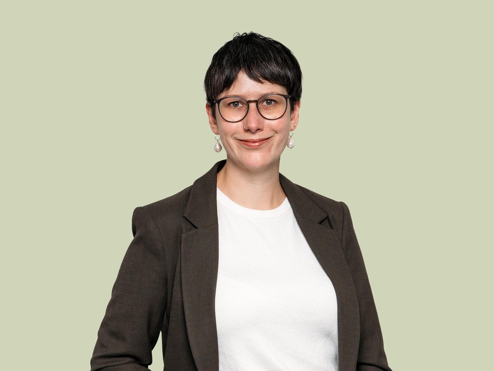 Dr. Verena Suchy, Person with short dark hair, glasses, and pearl earrings wearing a dark blazer and white shirt, smiling against a light green background.
