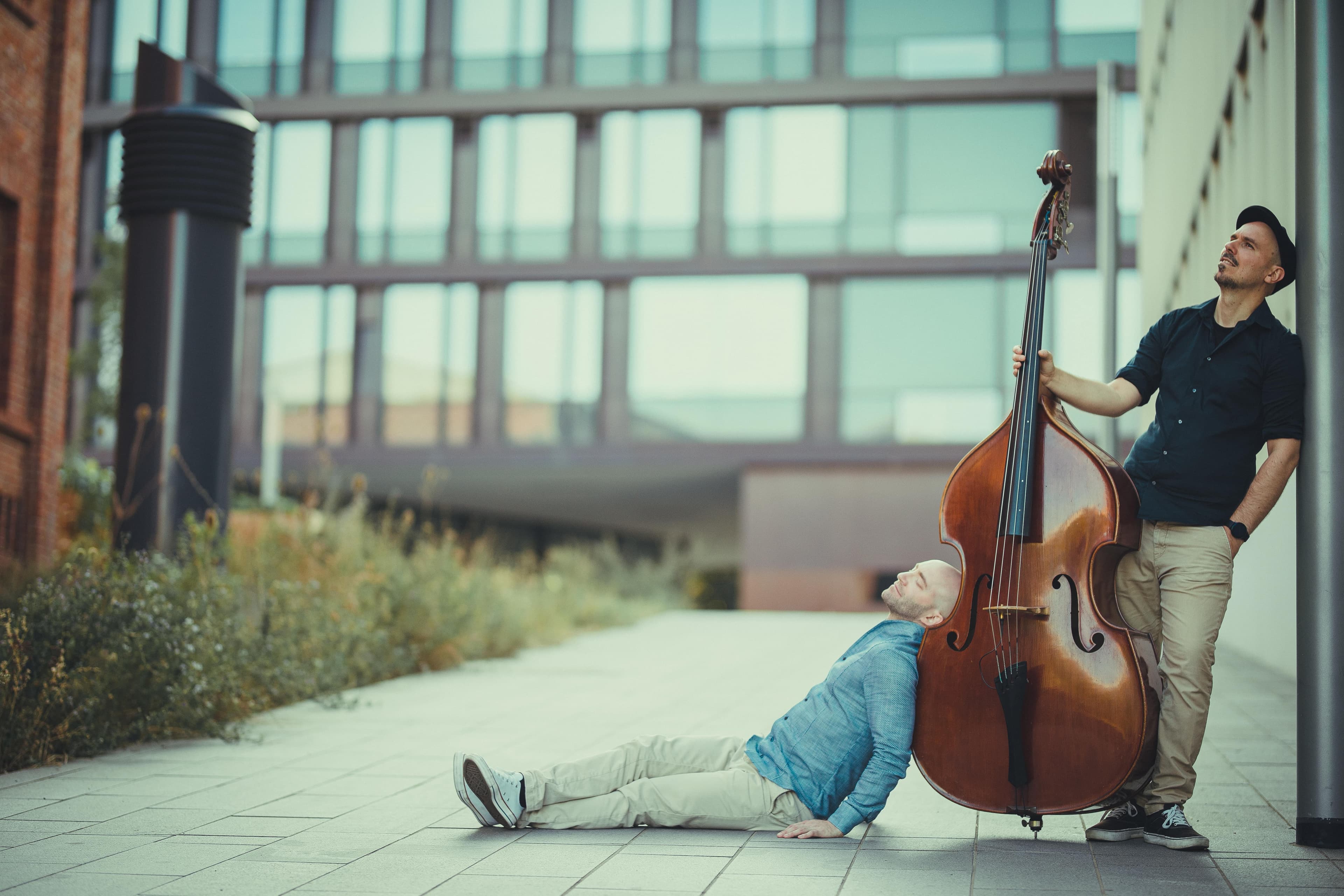 Two men outdoors, one sitting on the ground, the other standing and playing a double bass, with modern buildings in the background.