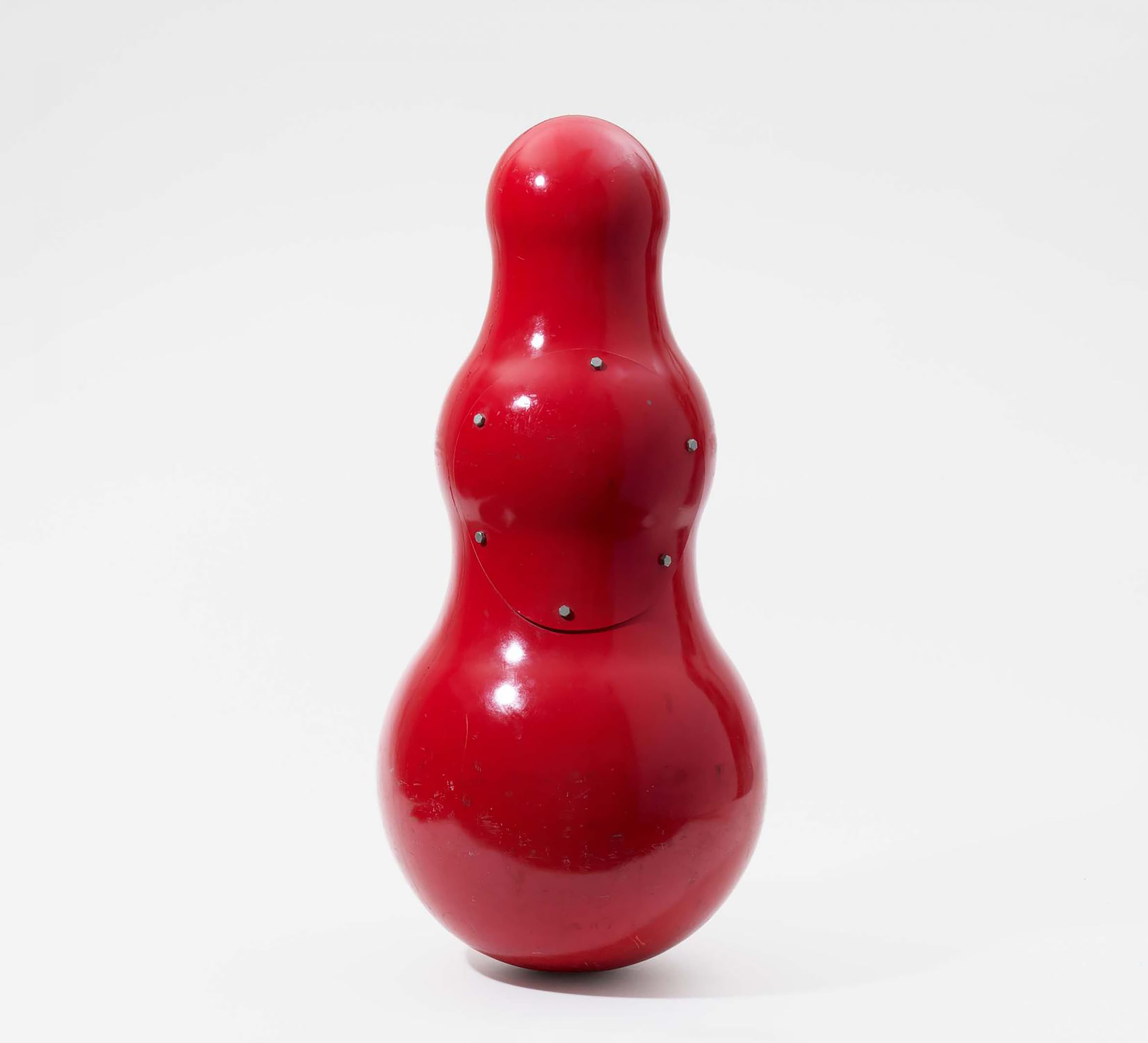 Rotes, glänzendes, kürbisförmiges Objekt mit glatter Oberfläche und kleinen, sichtbaren Schrauben. Steht aufrecht vor einem schlichten weißen Hintergrund.