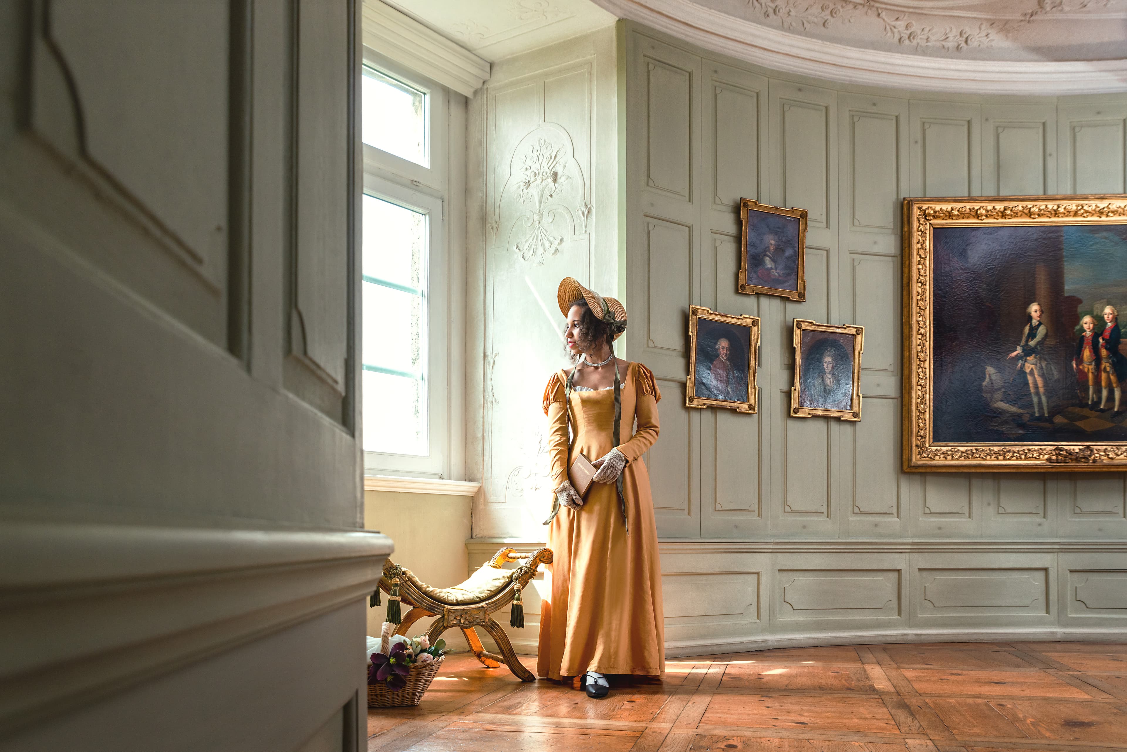 Eine Frau in einem langen gelben Kleid und Hut steht an einem Fenster in einem reich verzierten Raum mit Porträts an den Wänden und Holzboden.